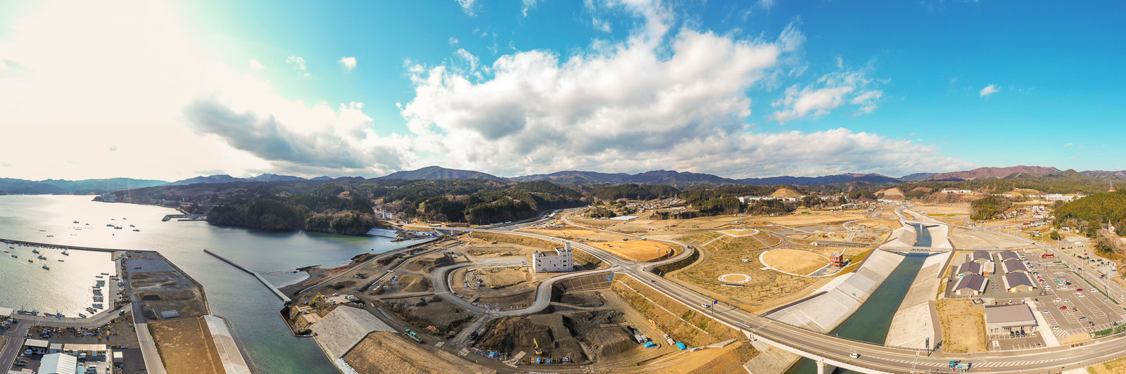 開発または再整備中の海岸地域を広く見渡した風景で、複数の建設現場や道路、建物が進行中で、左側には海岸線と港に停泊する船が見られ、背景には丘や山が広がり、空には雲が点在する海岸地域の写真