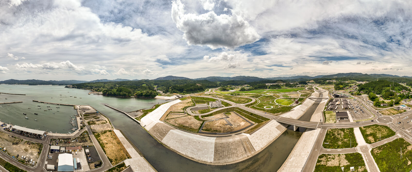 港湾の防波堤や川の堤防が整備され、周辺には整地された土地や道路が広がり、遠くに山々が見える昼間の風景を高台から定点カメラで撮影した写真
