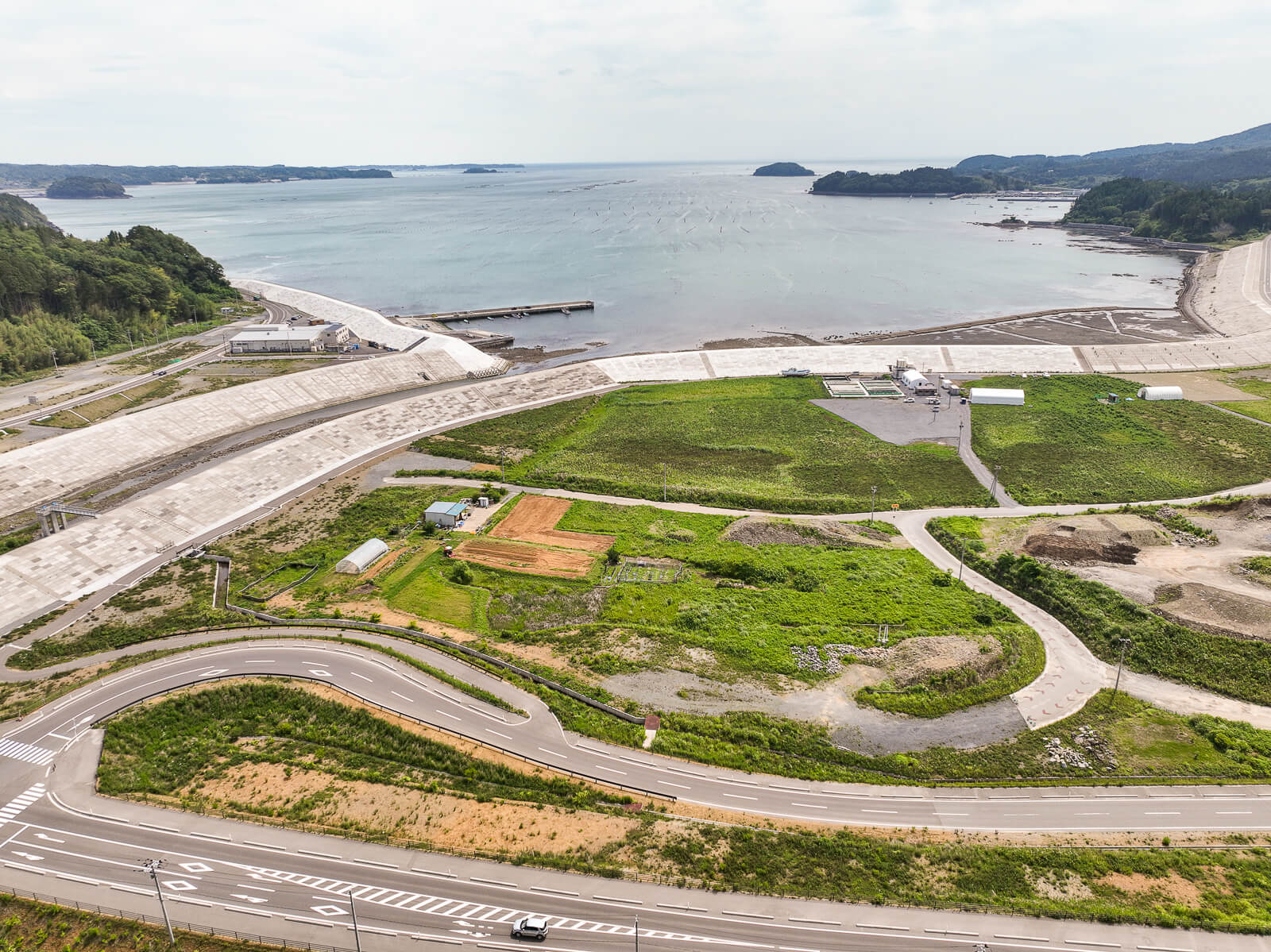 防潮堤の前に広がる緑地と畑、建物などが点在し、その先に湾の穏やかな海と防波堤が見える昼間の風景を高台から定点カメラで撮影した写真