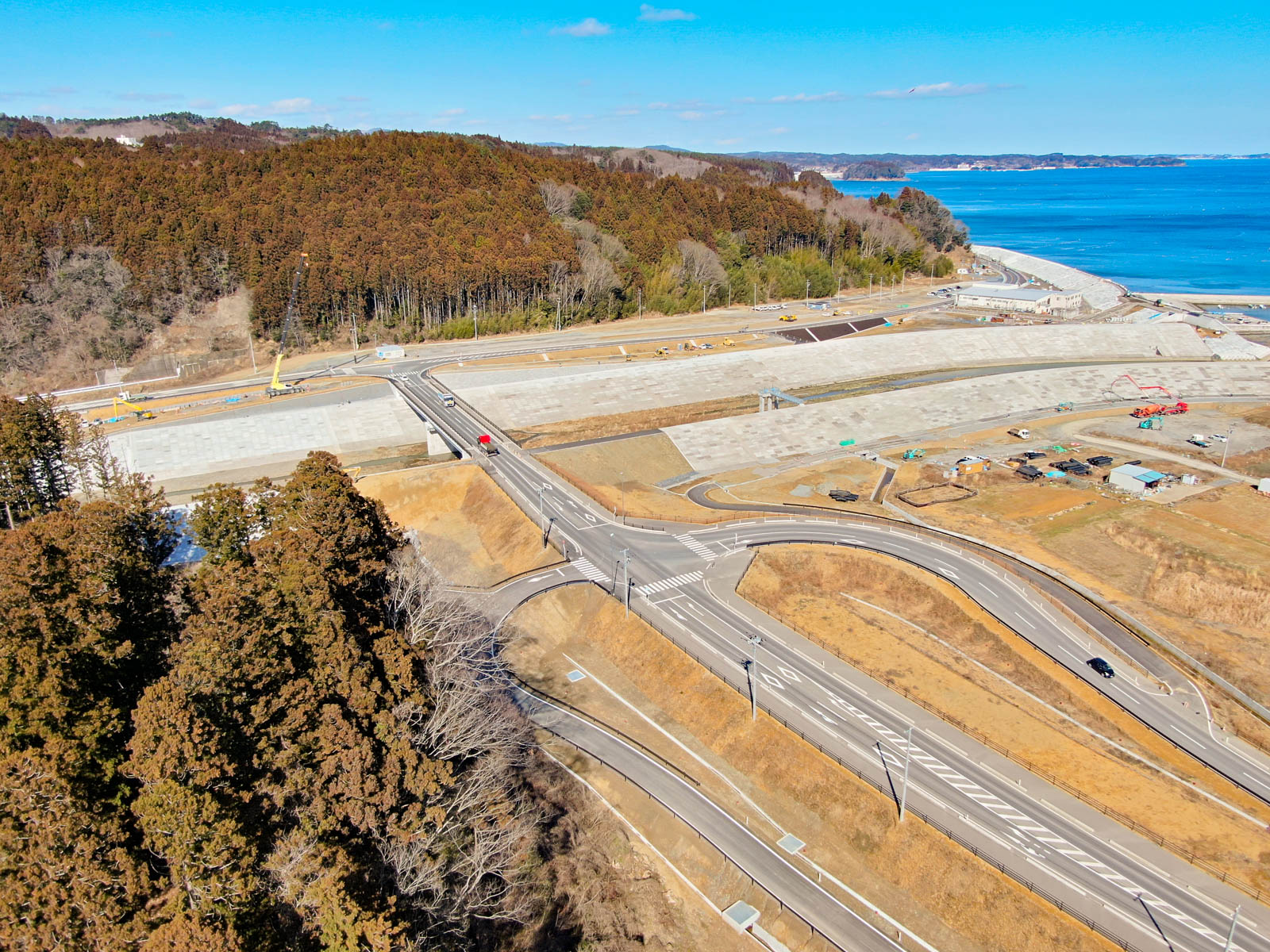 海岸地域を上空から見下ろした風景が広がっており、複数の道路や高速道路が網の目のように走り、橋がその一部を横断していて、道路上にはいくつかの車両が見え、左側には森林に覆われた丘陵地、右側には広がる海が写っている写真