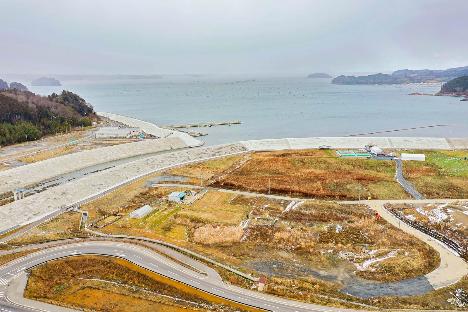 草地と畑が広がる手前に道路が曲がりくねって走り、奥にはコンクリートの防潮堤と広大な海が広がる、沿岸部の農地と防災インフラの昼間の風景を高台から定点カメラで撮影した写真