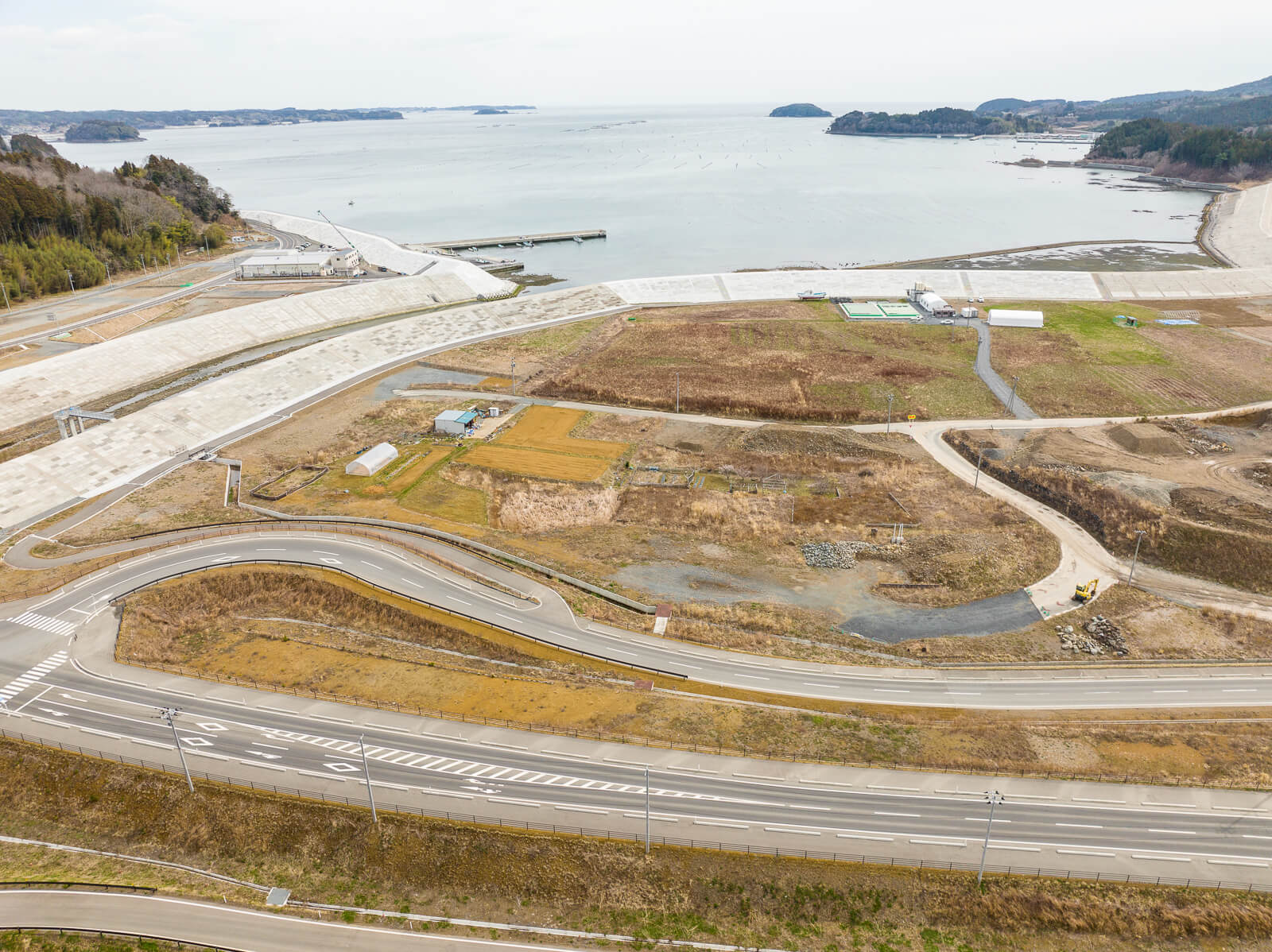 複数の防潮堤が重なるように続き、奥には静かな湾と島が見え、手前の道路はカーブしながら土地の起伏に沿って走っている、海辺の復興エリアの昼間の風景を高台から定点カメラで撮影した写真