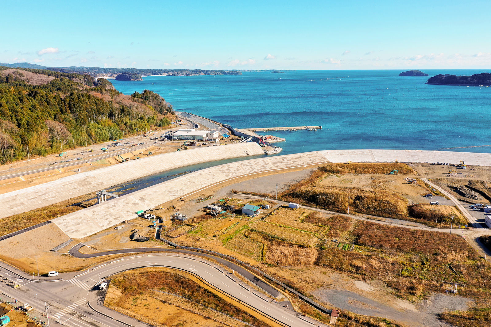 海岸沿いに広がる地域を上空から撮影した風景で、海に面して大きな防波堤または堤防が築かれており、その周辺には道路や建設機材、草地や開けた土地が点在し、海岸保護のための大規模なインフラ整備の様子が写された海岸地域の写真