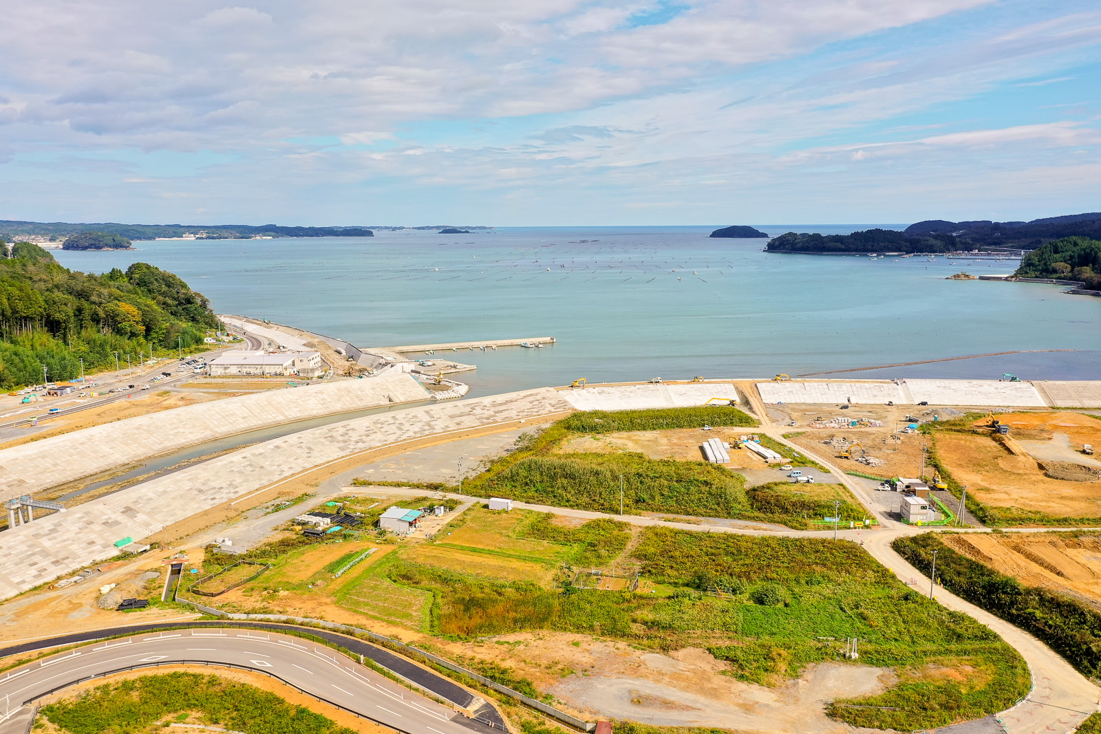 緑に覆われた土地と工事中の区域が入り混じる海沿いの地域で、手前には湾曲した道路が海岸へと続き、海には防波堤のような人工構造物が突き出し、空には青空と雲が広がっている海岸地域の写真