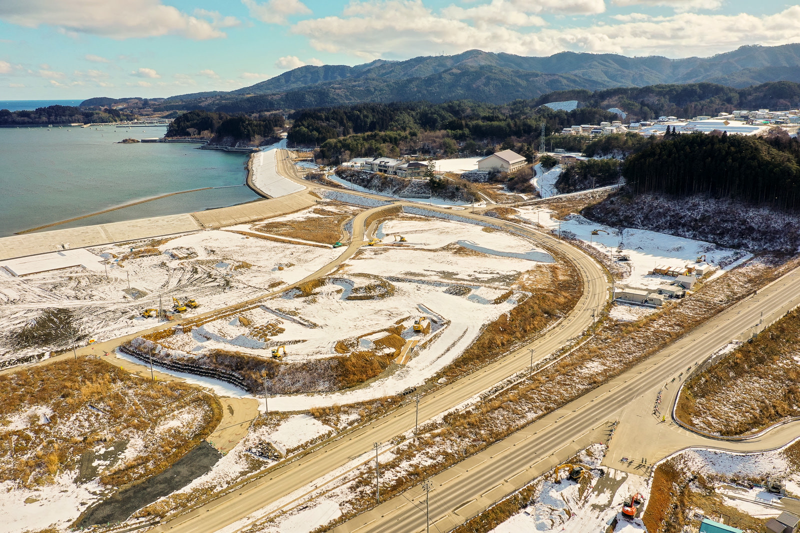 雪に覆われた海岸地域を上空から撮影した風景で、曲がりくねった道路が敷地内を走り、複数の建設車両や機材が点在しており、海岸には防波堤が築かれ、背景には山々が広がり、空には雲が浮かび、手前には海岸に沿って大きな道路が走る冬の海岸地域の写真