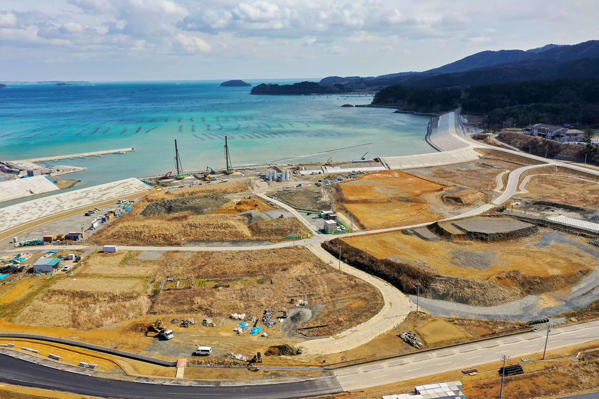 海岸沿いに広がる広大な建設現場で、重機や資材が点在し、地面は整地されており、背景には海が広がっている様子が写っている建設現場の写真