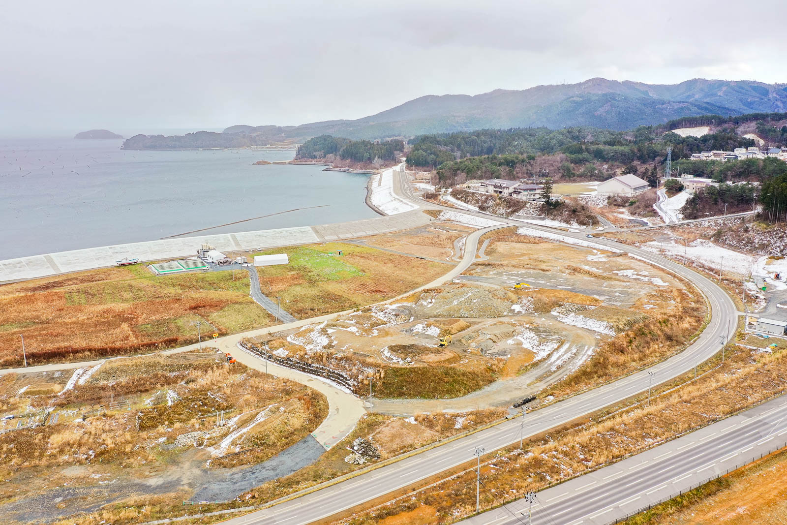 海沿いに防潮堤と小さな建物が見え、その奥には湾が広がり、遠くの山々まで見渡せる、湾岸地域の再開発中の昼間の風景を高台から定点カメラで撮影した写真