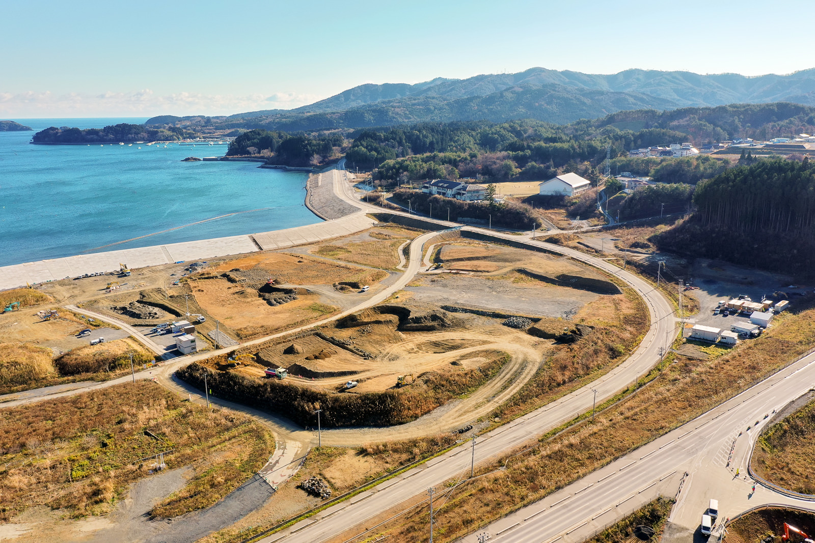 青く澄んだ海と砂浜が広がる海岸地域を高所から撮影した風景で、背景には森林に覆われた丘や山が連なり、手前には開発中と思われる土地に曲がりくねった道路や小道が走り、海岸近くには建物や構造物、道路沿いには車両も見られる広域の海岸地域の写真