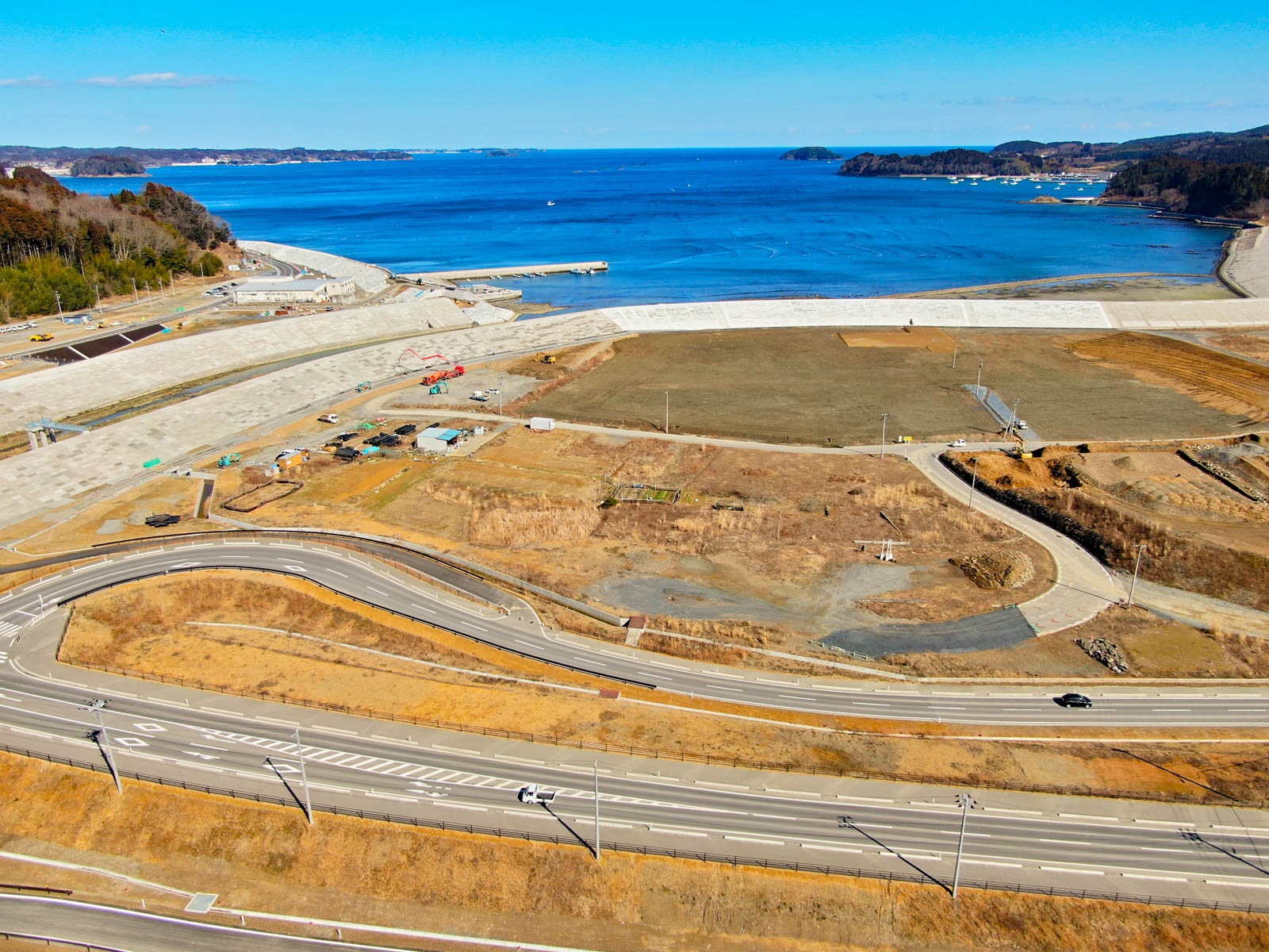 海を背景にした海岸地域が写っており、手前には曲がりくねった道路が走り、海岸近くの土地では建設または開発作業が行われている様子が見られ、草地や建物、複数の通路が点在し、空は澄んだ青空で天候が良好な写真