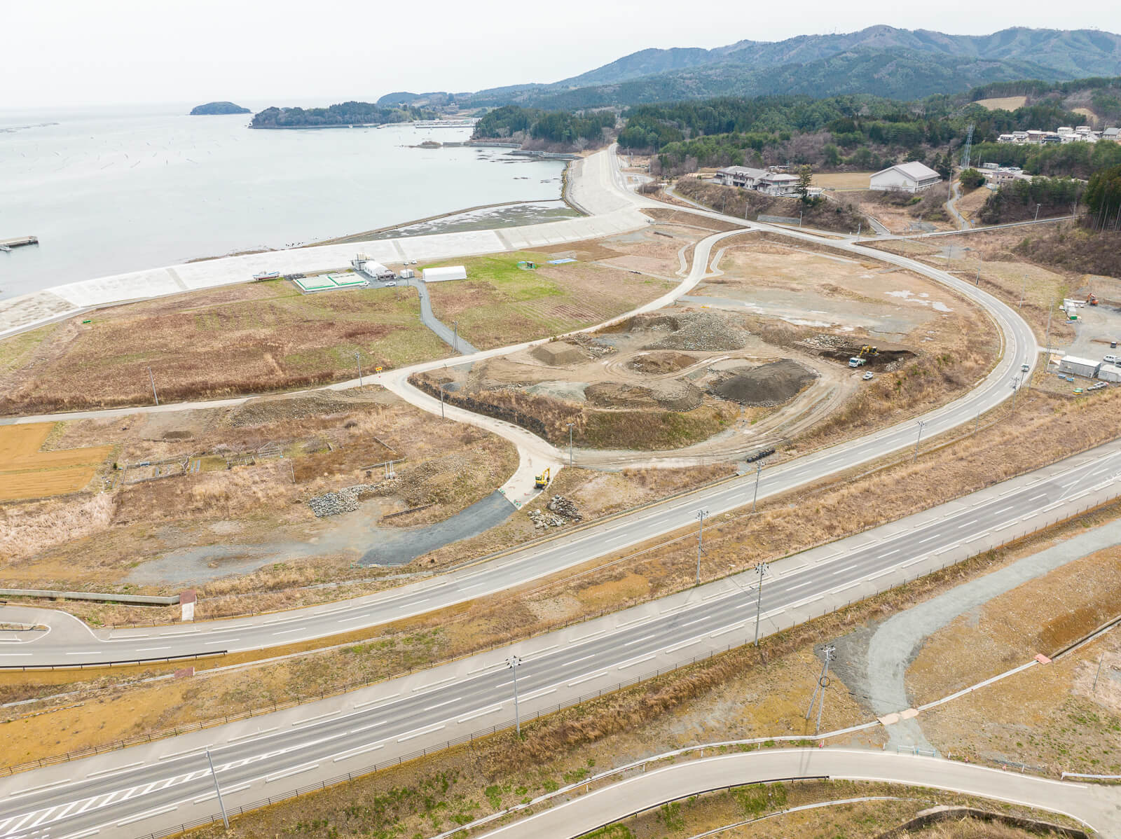 舗装された道路が蛇行しながら丘を登り、右手に造成中の土地区画や重機が見え、奥には湾と山が広がる、再開発中の沿岸地域の昼間の風景を高台から定点カメラで撮影した写真