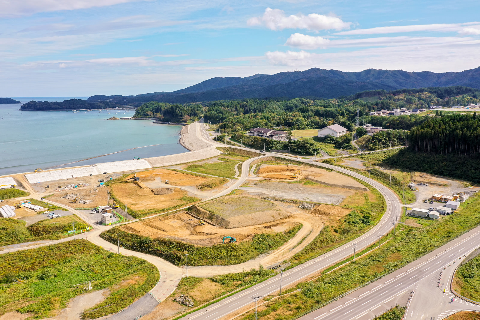 左側に海が広がり、コンクリートの防波堤で囲まれた沿岸部に、曲がりくねった道路や整地された土地、建設機材が点在し、背景には森林に覆われた丘と建物が散在する様子が空撮で捉えられた沿岸開発地域の写真