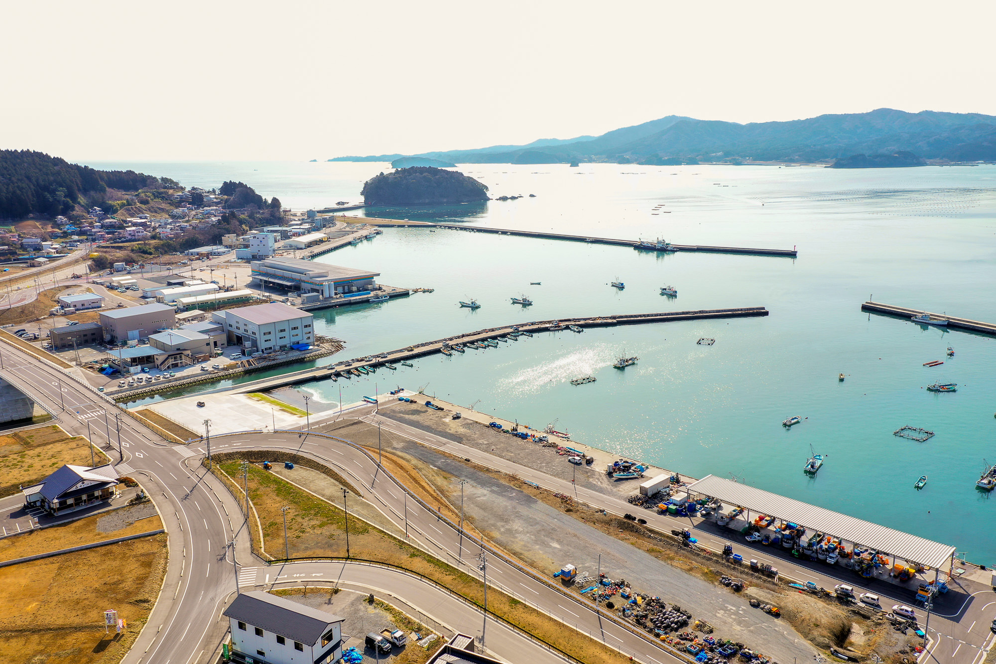 湾に面した港町を空から見下ろし、複数の桟橋に船が停泊し、海沿いには倉庫のような建物が並び、町は海岸線に沿って湾曲した道路に沿って広がり、背景には山々と遠くに島が見える穏やかな海辺の町の写真