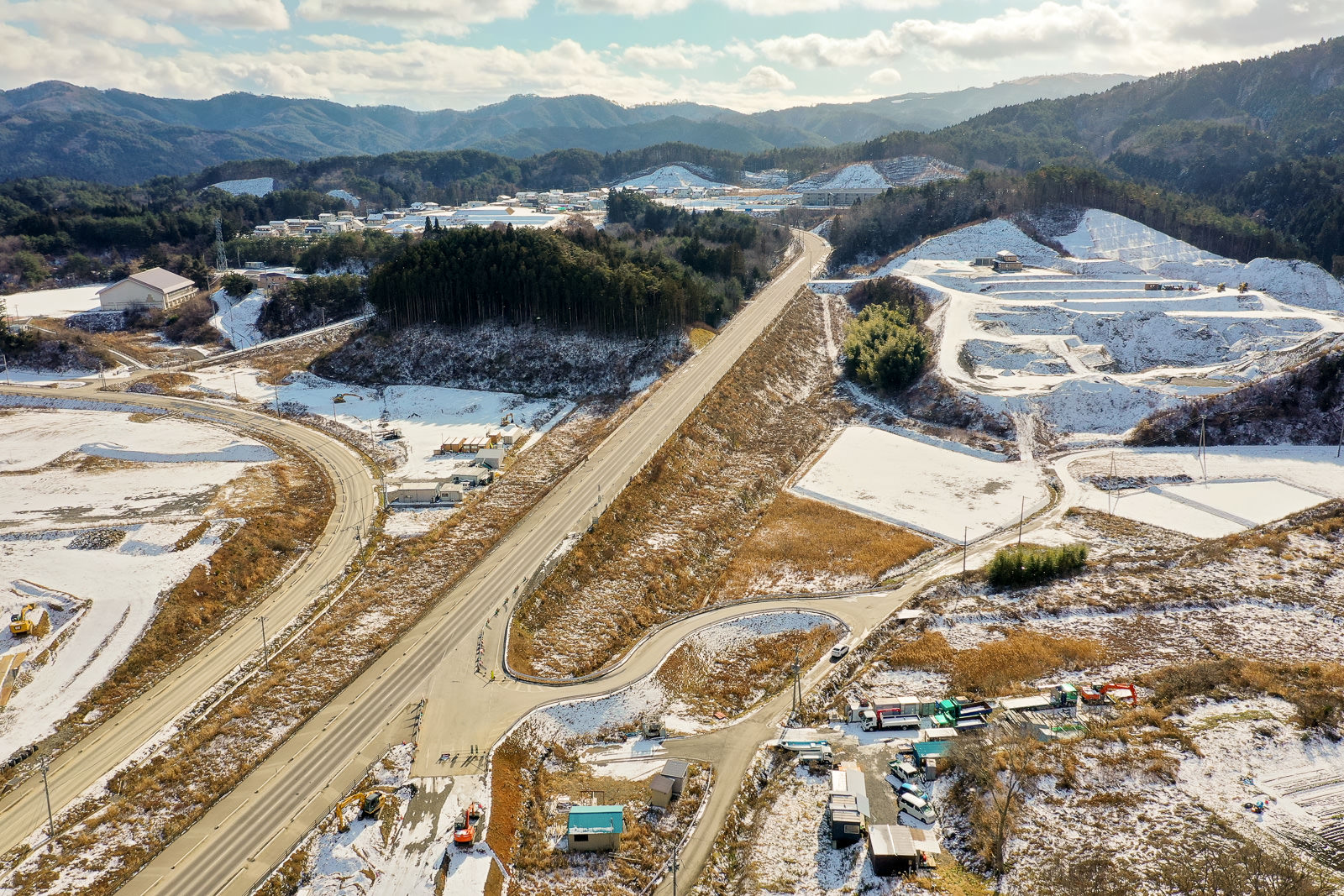 冬の農村地域を上空から撮影した風景で、中央には幹線道路が走り、そこから枝分かれする小道が広がり、地面には雪が部分的に積もり、複数の建物や構造物が点在し、背景には木々に覆われた丘や山が広がり、空には雲が浮かびつつも日差しが差し込む冬の写真