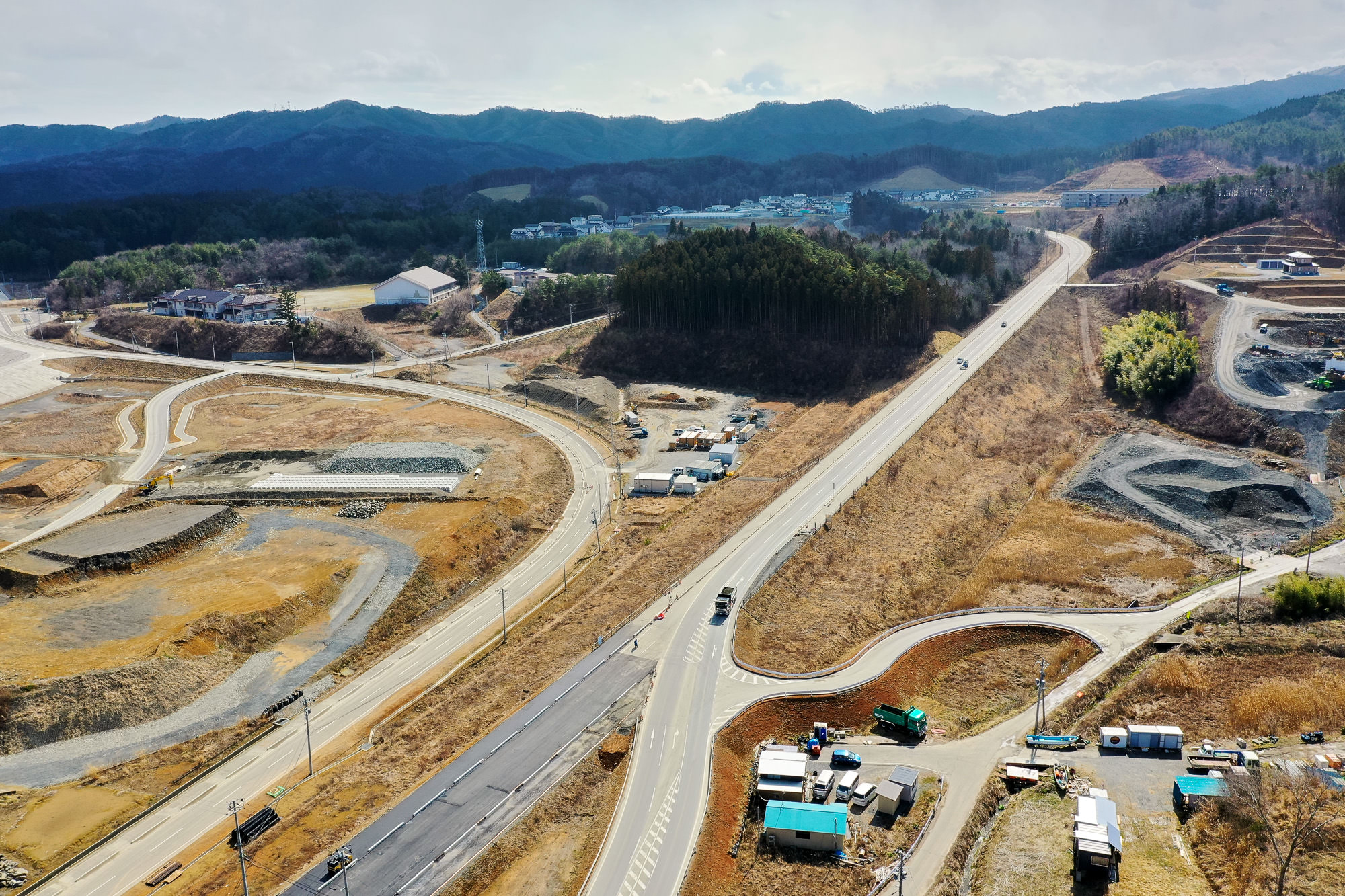 中央に幹線道路が通り、そこから複数の小道が枝分かれし、周囲には建物や構造物が点在し、背景には丘や山が広がっている、自然の地形と整備された土地が混在する風景の写真