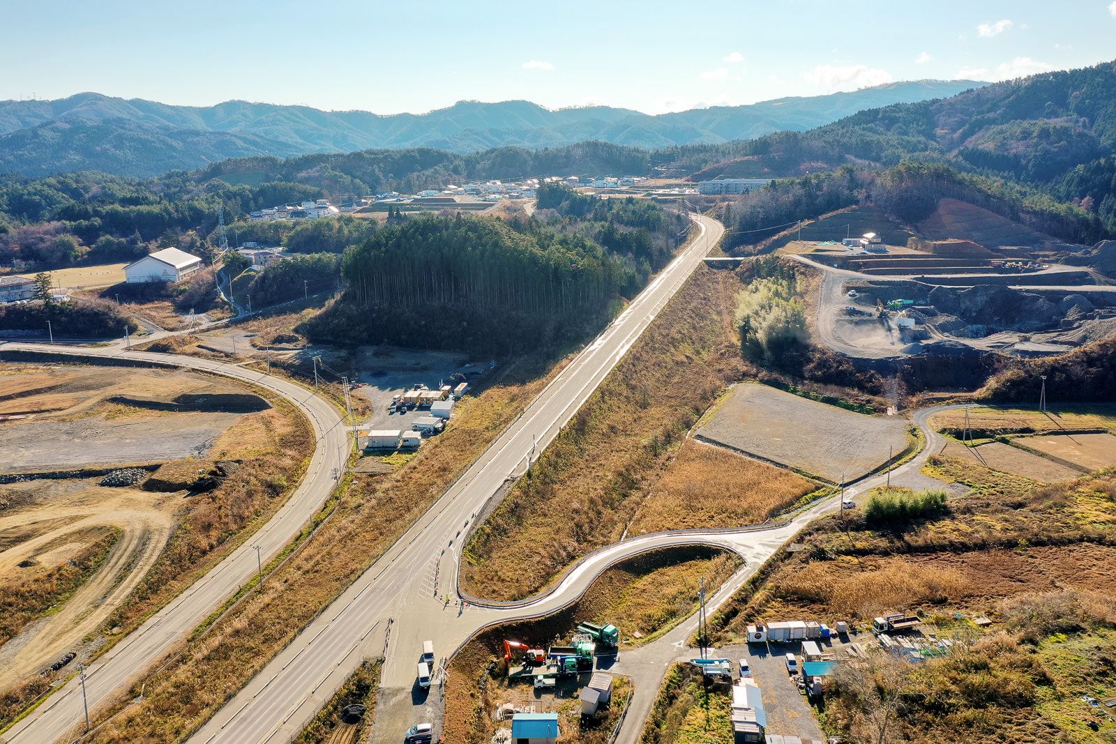 田園風景の中を曲がりくねった道路が走っており、その道路は新しく造られたか建設中で、画像の下部には工事用車両や機材が見られ、周囲には畑や建物、背景には森林に覆われた丘が広がる農村地域の写真