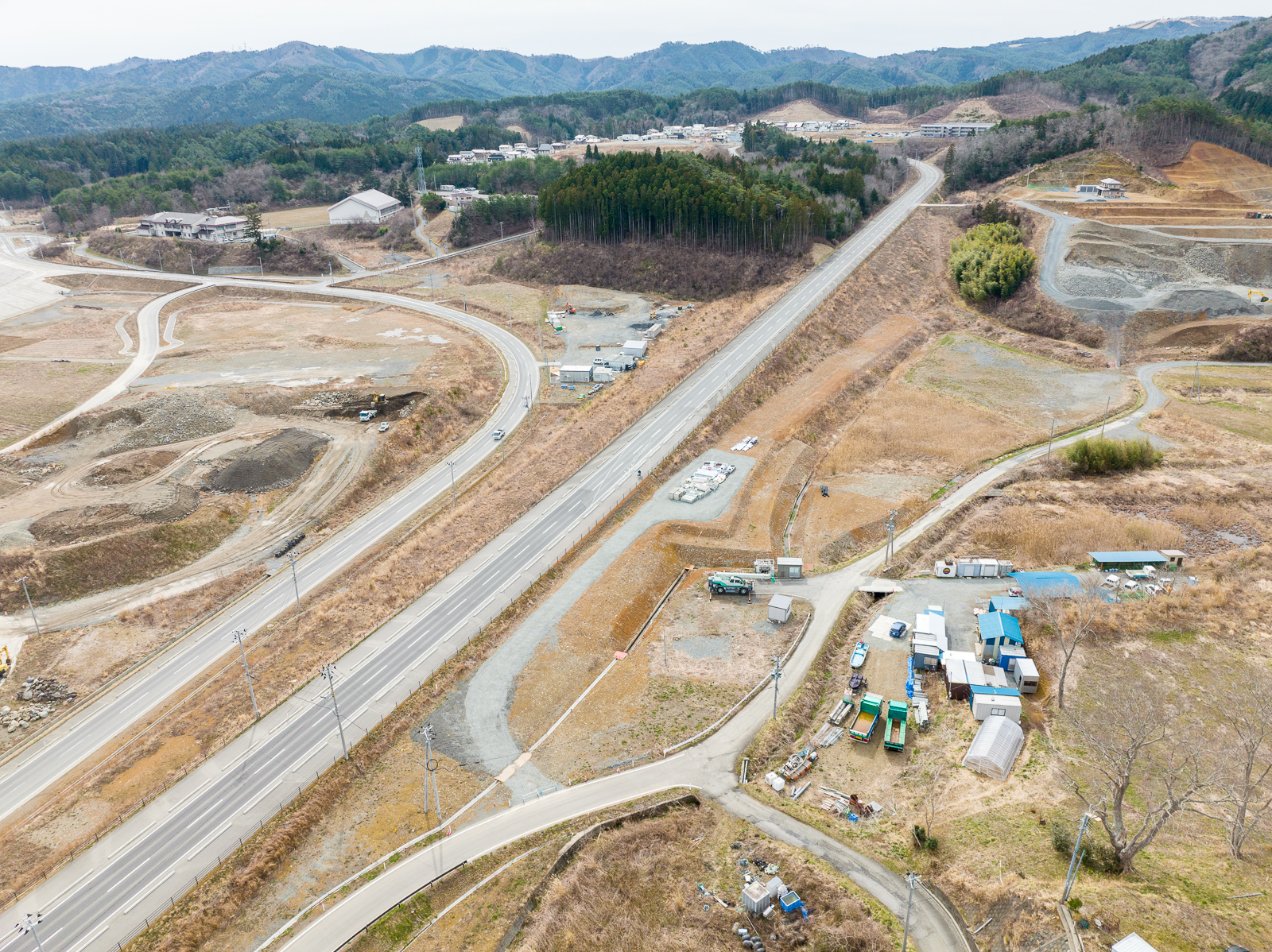 山間部の麓から高台へ伸びる幹線道路沿いに、資材置き場や仮設建物、小規模な作業場が点在しており、遠くには住宅地が広がる、復興インフラの終点部の昼間の風景を高台から定点カメラで撮影した写真