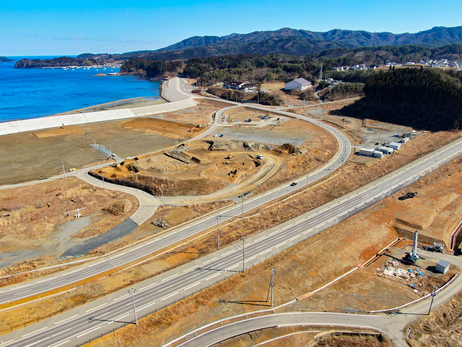 海岸地域の風景が広がっており、左側には海が陸地に接していて、中央には建設現場と機材が点在し、複数の道路が走っていて、幹線道路と小さな接続道路が交差し、背景には緑に覆われた丘や山々が広がり、空は澄んだ青空が広がっている写真