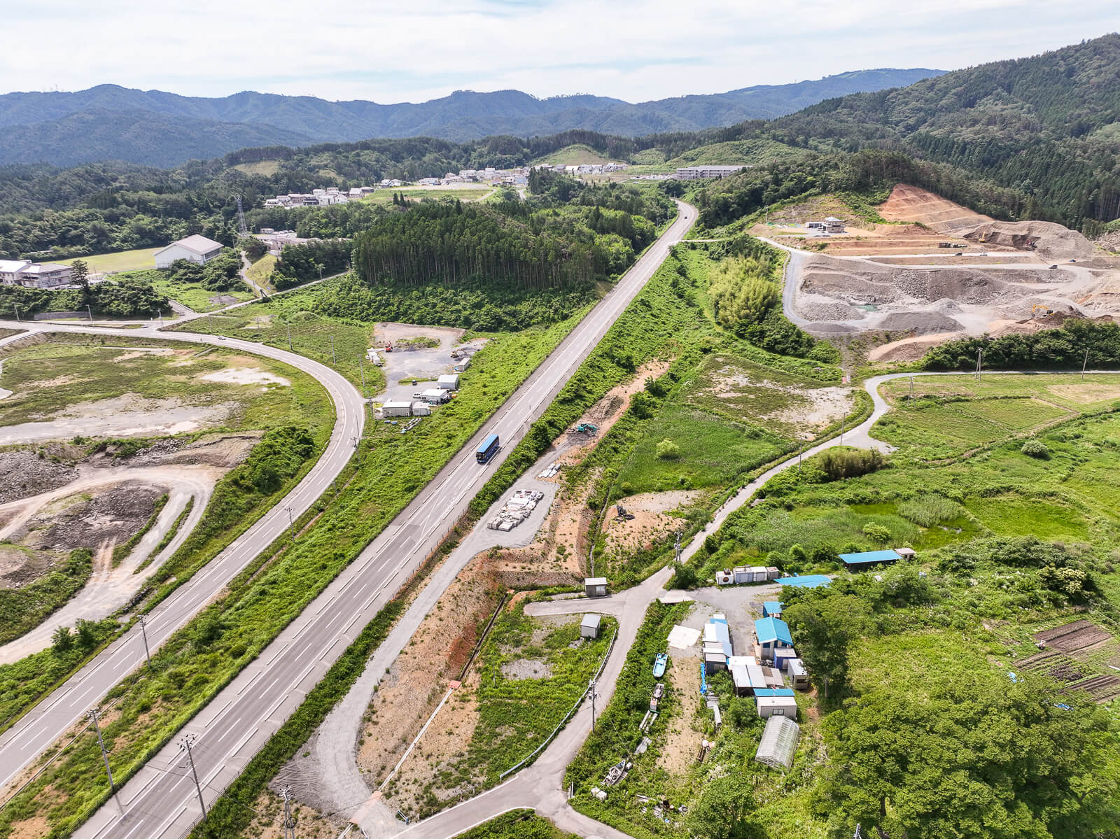 丘の中腹にある採石場や集落と、それに向かって伸びる道路、その両側に緑が生い茂る農地や山林が広がる昼間の風景を高台から定点カメラで撮影した写真