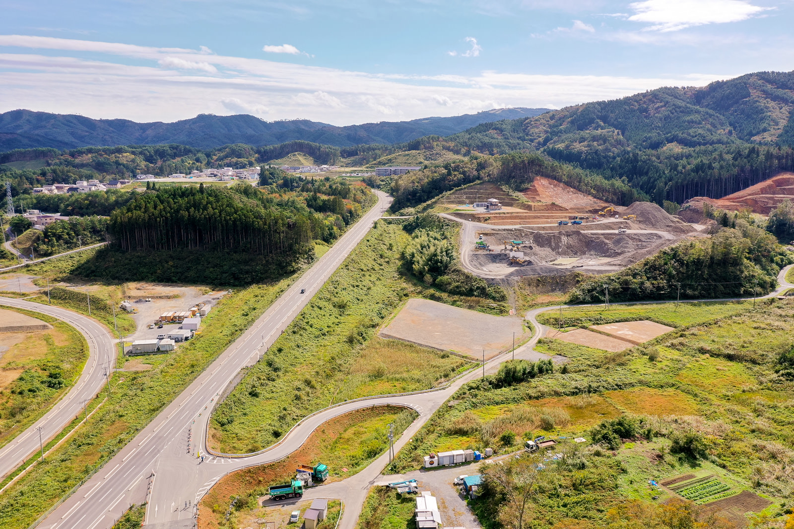 田園風景を空から見下ろした構図で、中央を道路が貫き、右側には重機が並ぶ工事現場が広がり、周囲には森林や丘陵、点在する建物と農地が見える晴天の農村地域の写真