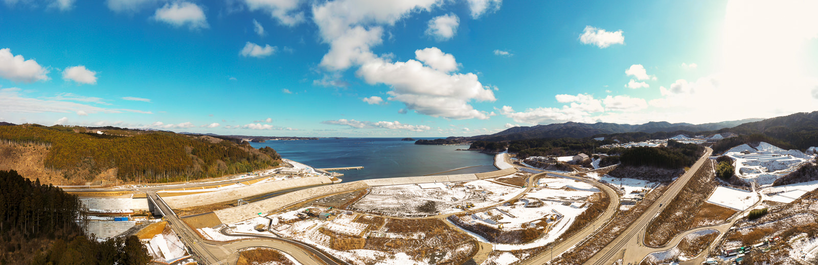 海を中心とした広大な海岸風景を上空から撮影した画像で、背景には大きな水面が広がり、周囲の丘陵地には雪が点在し、冬の季節を感じさせる景色で、手前には橋や曲がりくねった道路などのインフラが整備され、空は晴れて雲が少し浮かぶ冬の海岸地域の写真