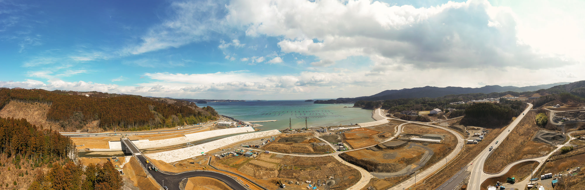 自然と開発が混在する海沿いの地域を広く見渡した写真で、背景には広がる海と海に突き出た構造物があり、手前には複数の工事現場や道路、造成中の土地が点在し、周囲には木々に覆われた丘が広がり、空には青空と雲が見える写真