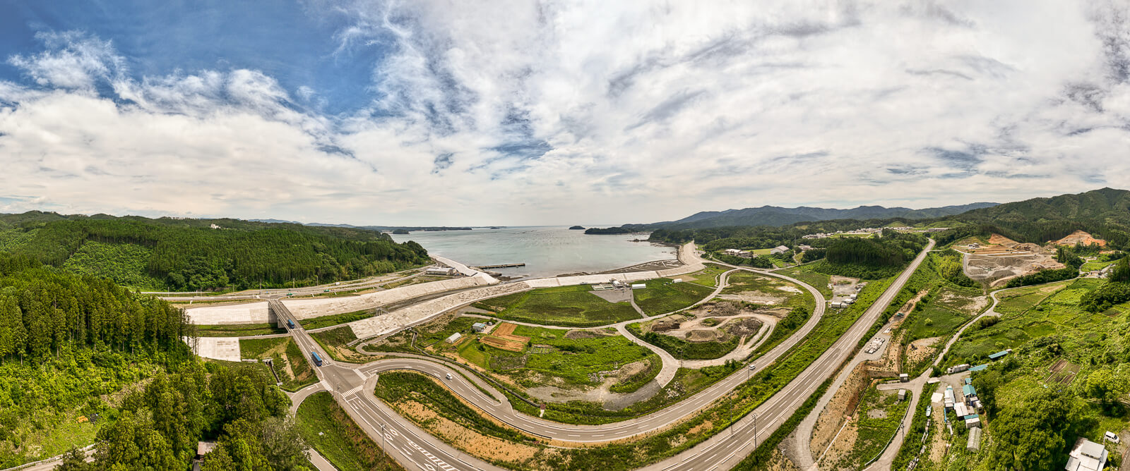 海に面した防潮堤の前に広がる草地と小規模な農地や施設が見え、背後には森や山が広がっている昼間の風景を高台から定点カメラで撮影した写真