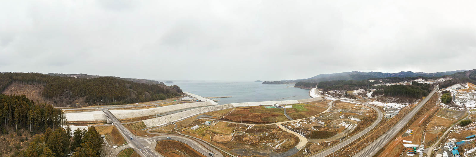 防潮堤で囲まれた広大な更地と道路が海岸線に沿って広がり、その奥には穏やかな海と湾が見え、全体に雪がうっすら積もっている沿岸部の再開発区域の昼間の風景を高台から定点カメラで撮影した写真