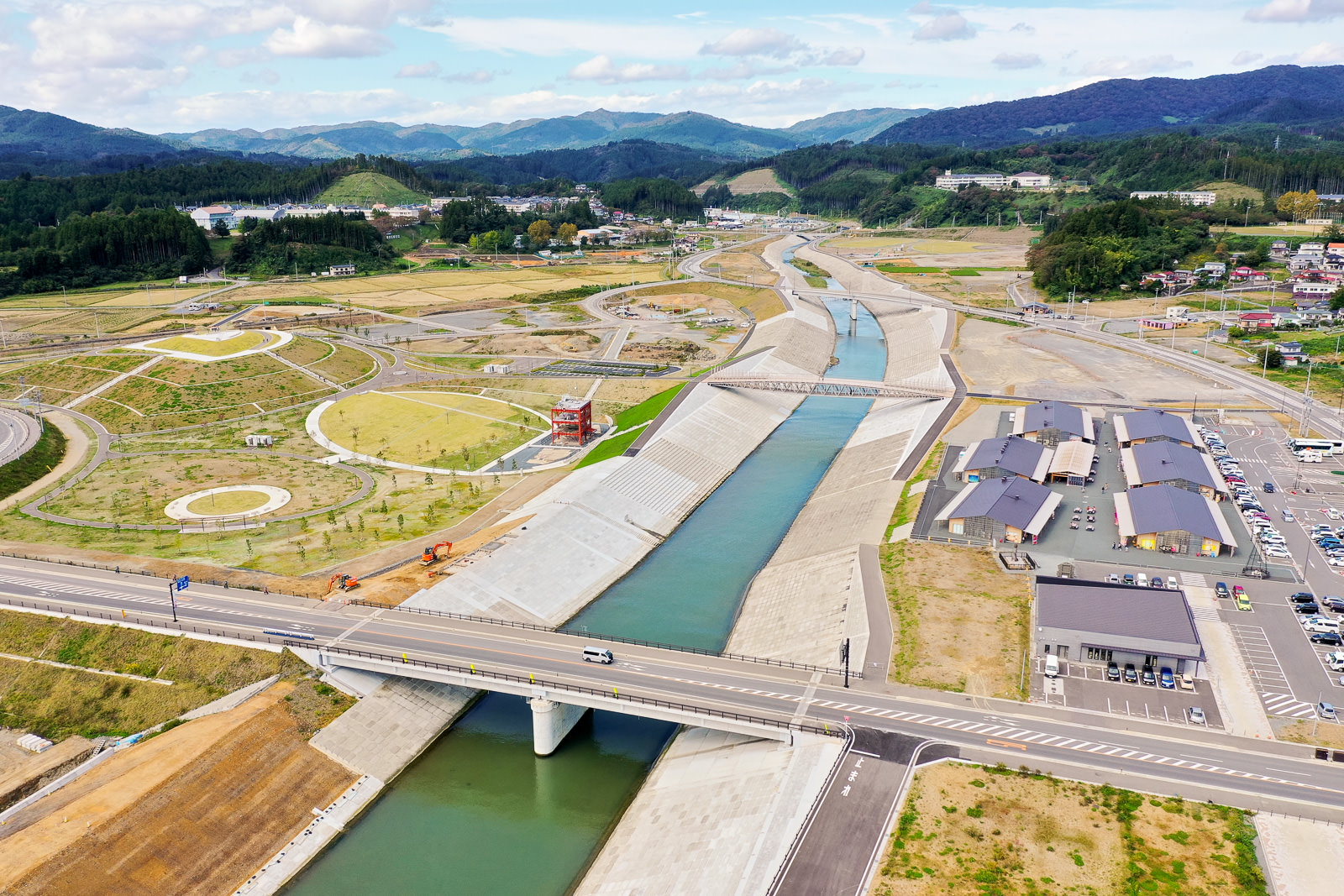 コンクリートの水路を流れる川を中心に、緑の田畑や丘陵に囲まれた地域を空から捉え、手前には橋が川を横断し、両岸には道路や建物、駐車場や歩道などのインフラが整備された開発中または開発済みの地域が広がる風景の写真