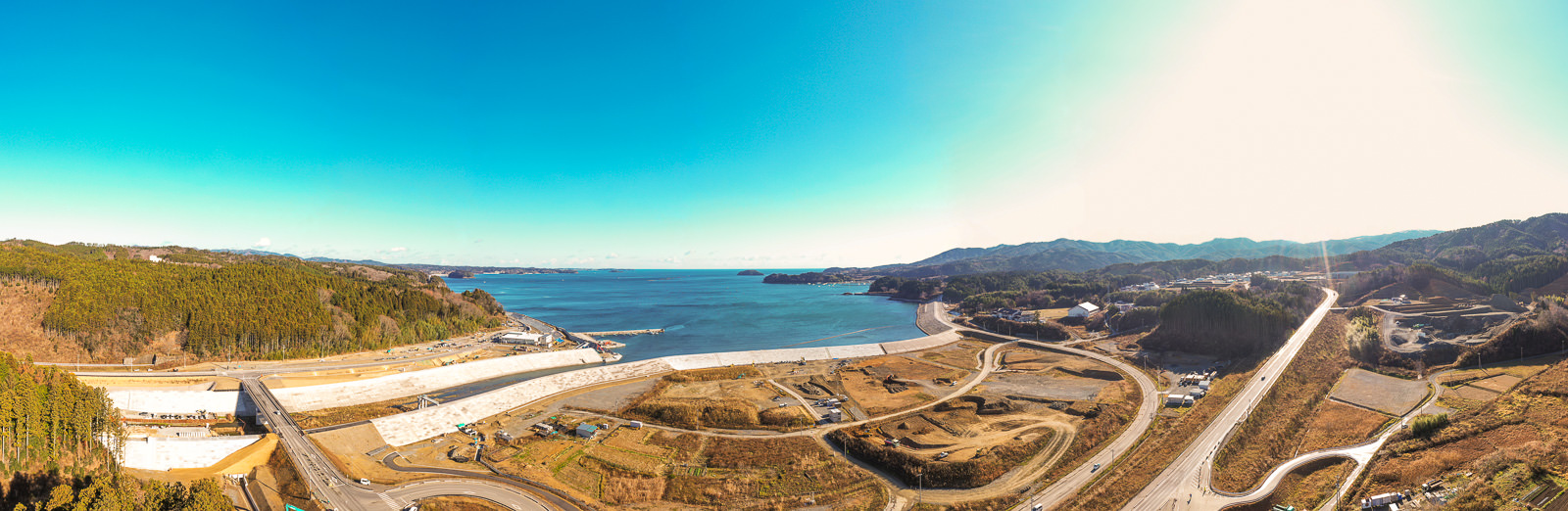 海に面した広大な沿岸地域を一望できる風景で、手前には道路や小道が入り組み、建設中の場所や草木が点在し、左側には丘と森林、右側には建物が点在する開けた土地が広がり、晴天の空から右側から日差しが差し込む様子が写された沿岸地域の写真