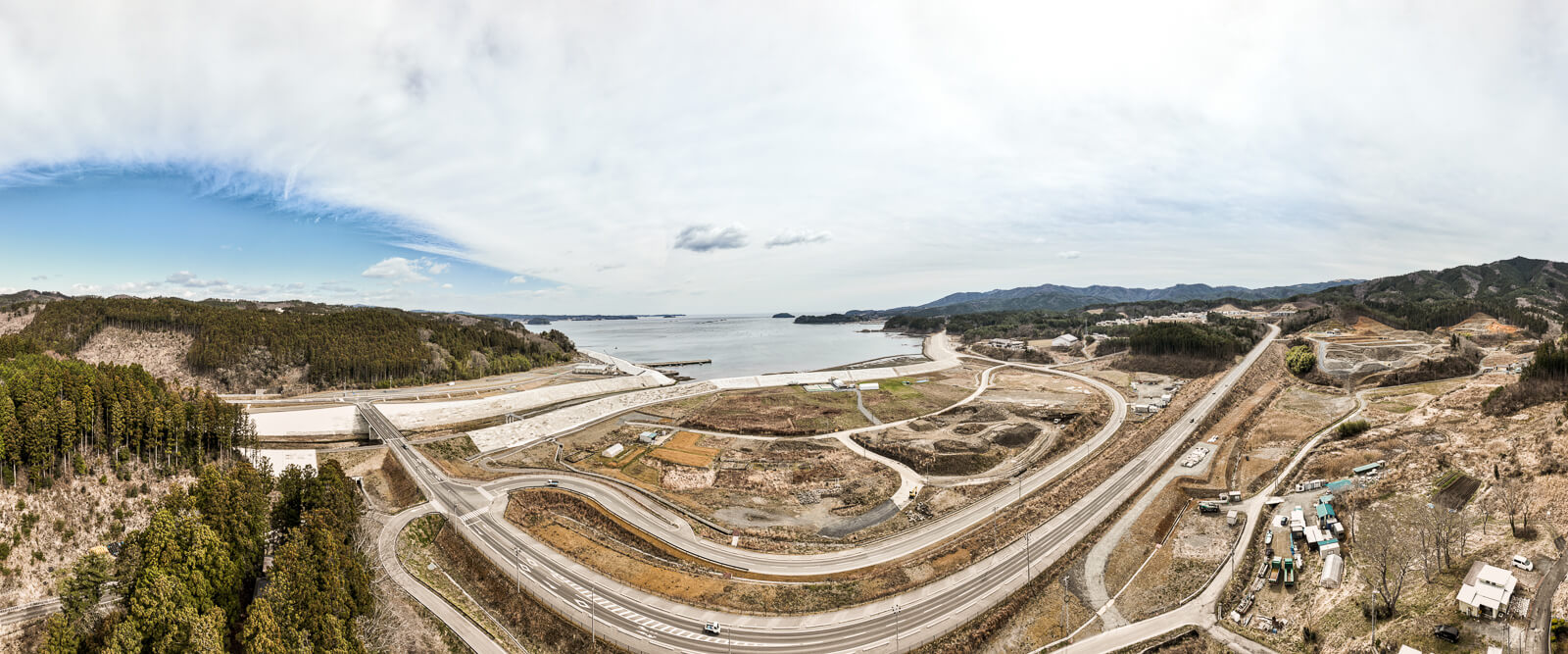 奥には海と防潮堤、手前には新しい道路が曲線を描いて走る風景が広がっている、海岸部の再整備中の昼間の風景を高台から定点カメラで撮影した写真
