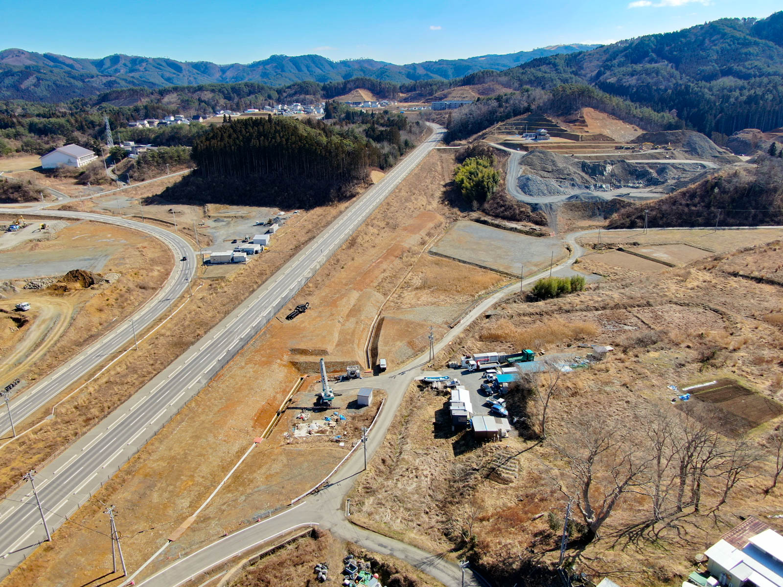 山間部の農村地域を上空から見下ろした風景が広がっており、中央にはまっすぐに伸びる幹線道路が走り、そこから複数の小さな道路が分岐していて、周囲には森林に覆われた丘陵地が広がり、道路付近には建物や建設機材が点在し、土地の一部は開発中で整地や機械が見える写真