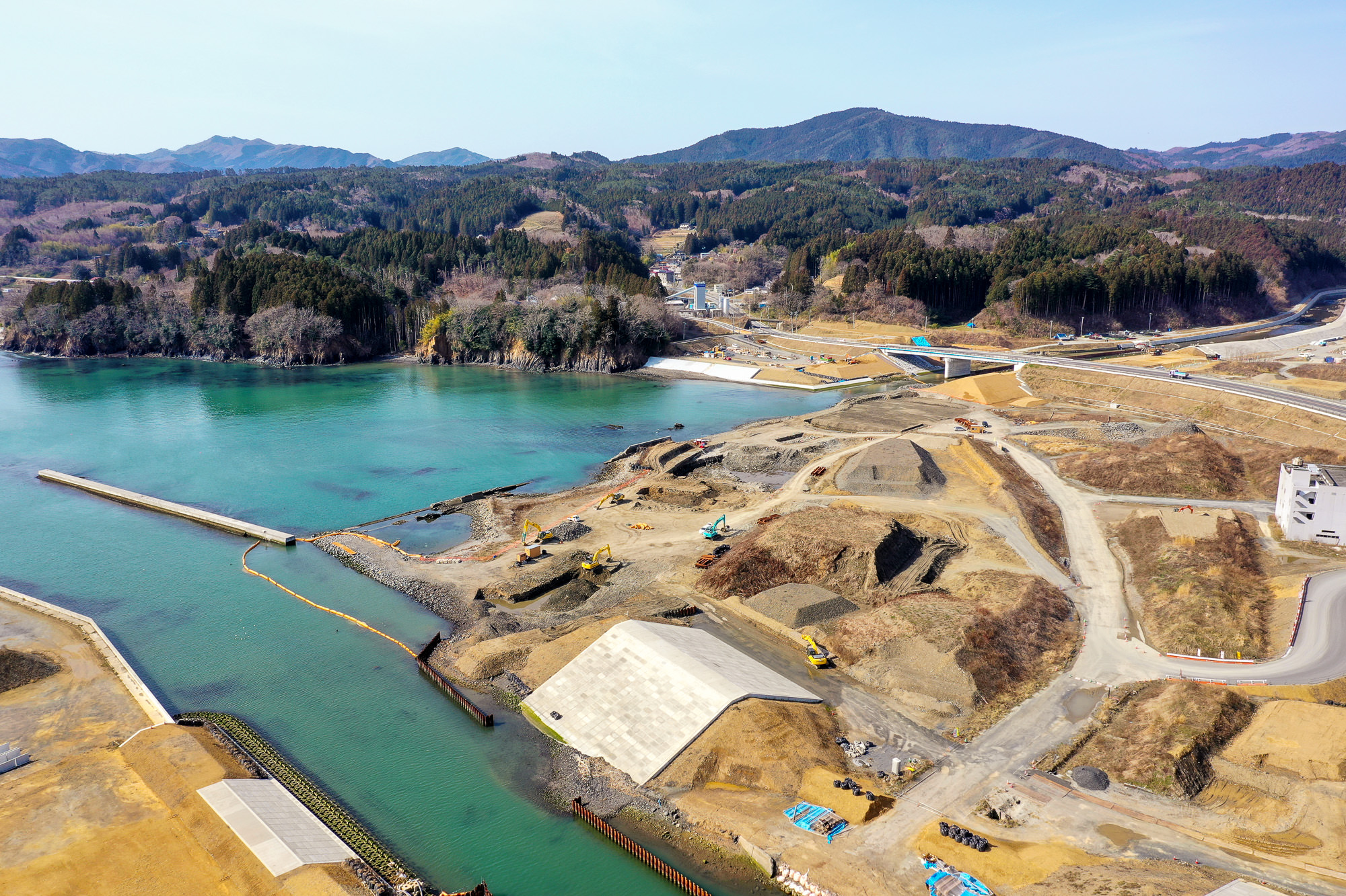 海岸沿いの工事現場を高所から見下ろし、整備中の土地の向こうに山々と森林に囲まれた穏やかな湾が広がっている様子を写した海辺の工事現場の写真