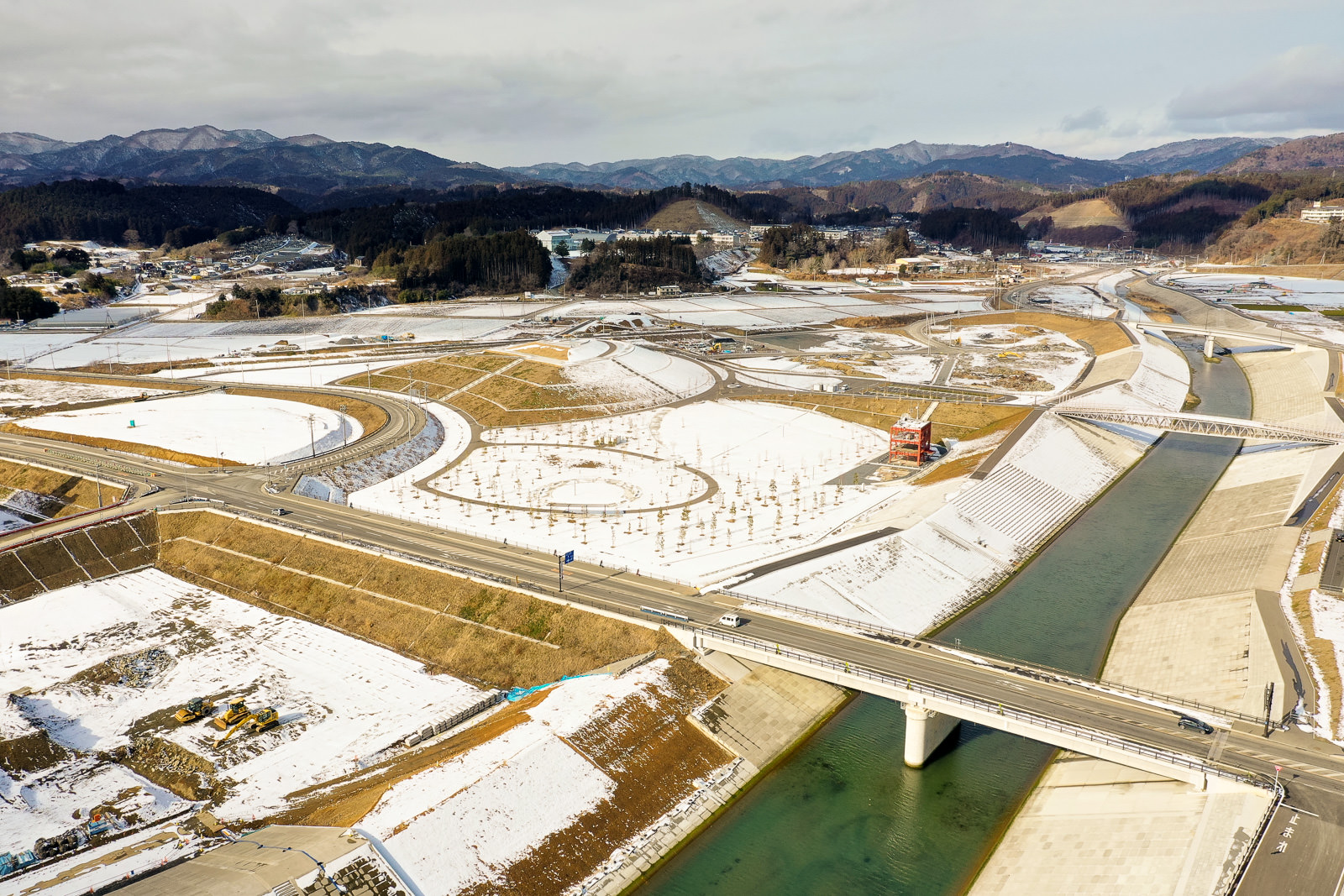 雪が積もった広い田園地帯の中に、川が流れ、その上に橋が架かっている様子を写した冬の風景の写真