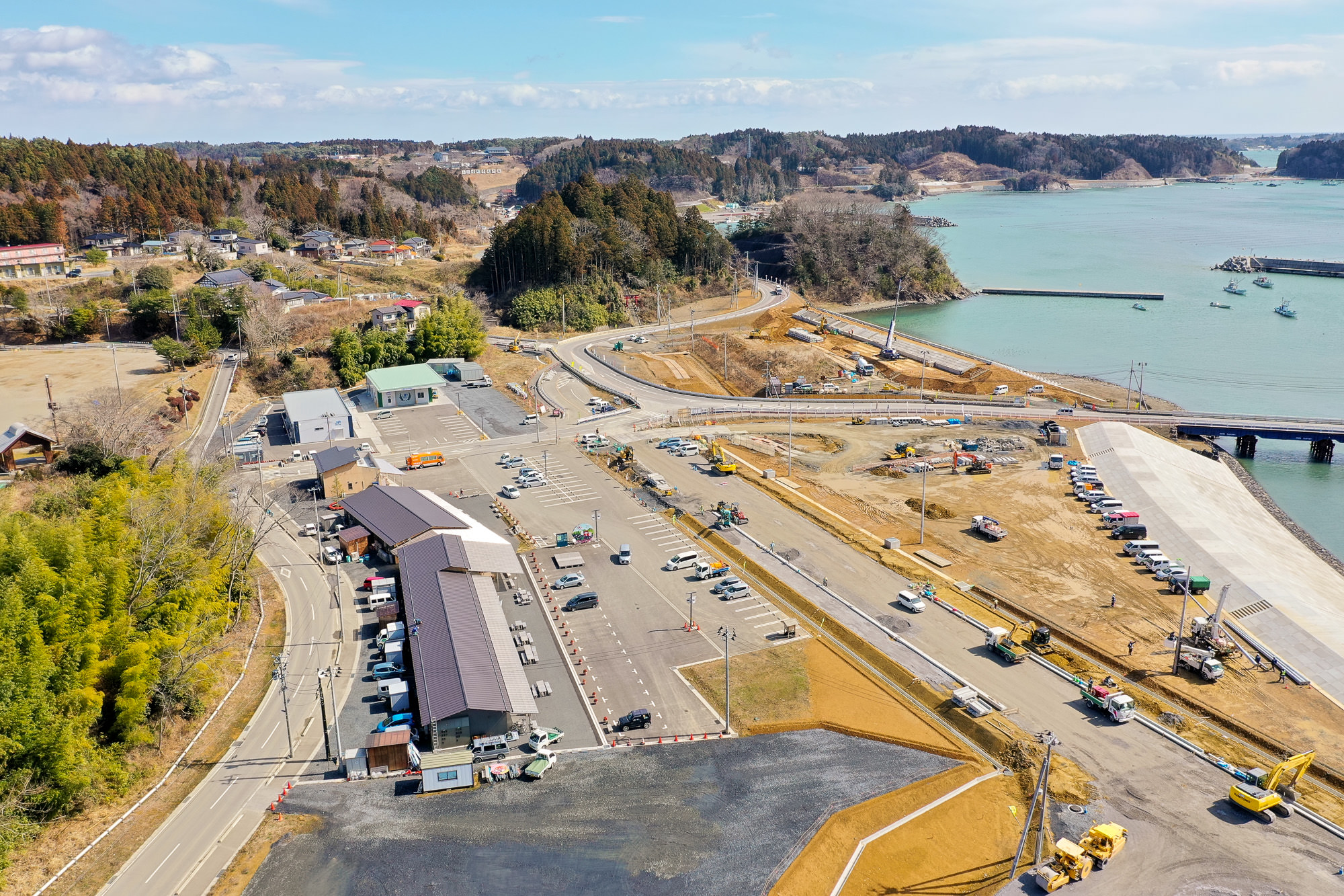 右側に海岸線が広がり、海には数隻の船が浮かび、陸地には複数の道路や建物、車両が見られ、背景には木々と丘が広がる開発中の沿岸地域を空から見下ろした海辺の開発地の写真