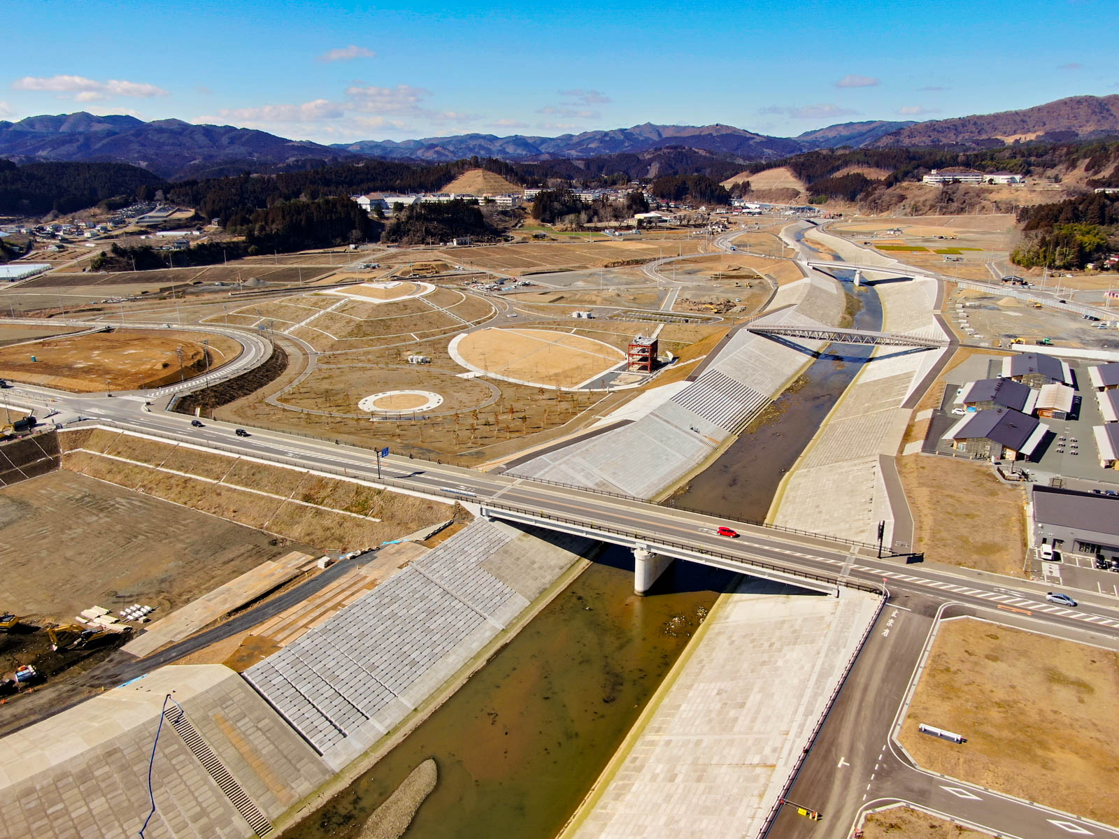 川に架かる橋を中心に、整然と配置された道路や通路が広がる風景が上空から写されており、円形や長方形の区画がいくつも見られ、将来的な開発や造園のための土地と考えられ、背景には丘や山々が広がり、右側にはいくつかの建物が見え、空にはわずかに雲が浮かぶ晴天の写真
