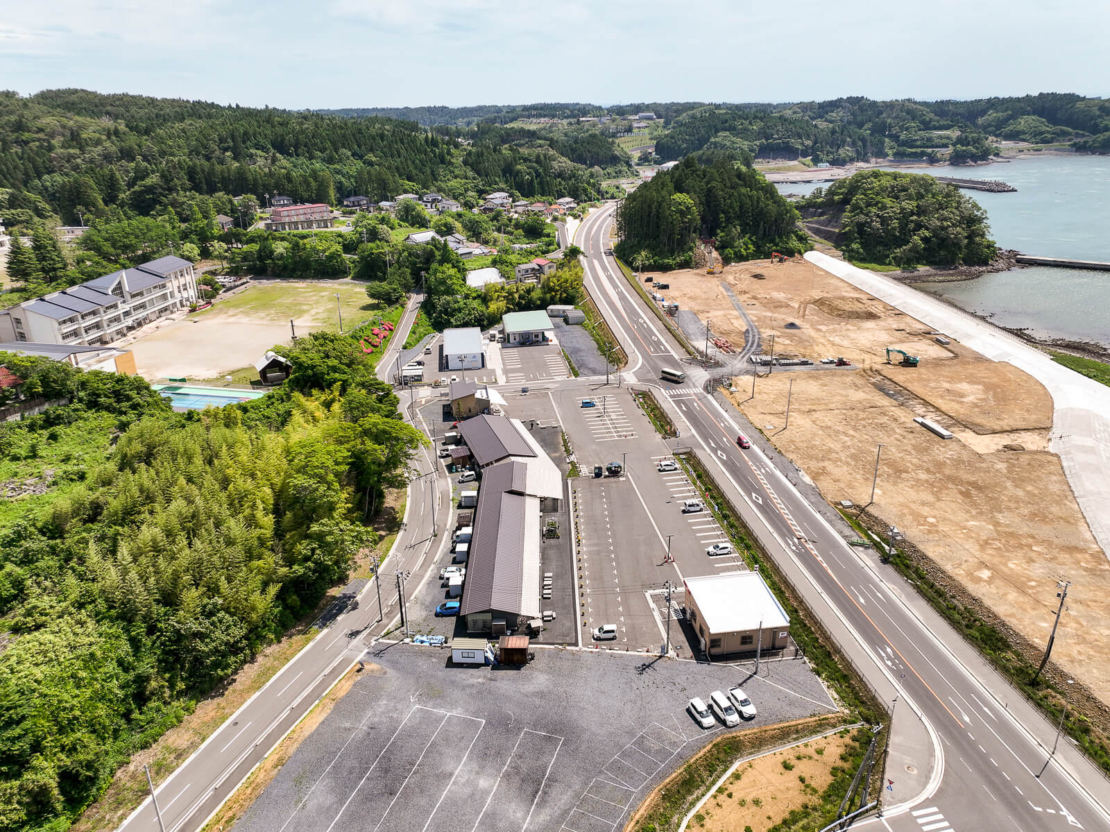 海岸線に沿って走る道路の横に駐車場や建物が並び、その向こう側の校舎の周囲に広がる緑豊かな山林と海が見える昼間の風景を高台から定点カメラで撮影した写真