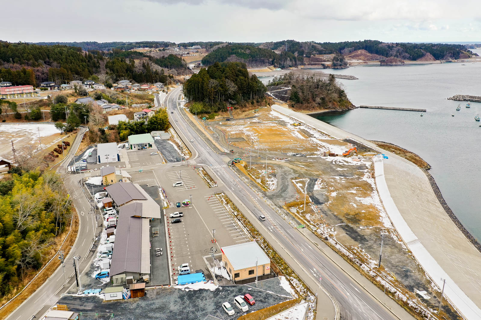 右側に海が広がり、中央には造成中の土地と重機が見え、左側には駐車場と複数の建物が立ち並び、奥には集落と森が広がる海沿いの再整備地域の昼間の風景を高台から定点カメラで撮影した写真