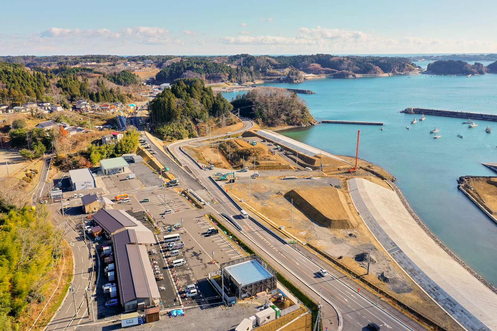 自然と開発が混在する海岸地域を上空から撮影した風景で、右側には水辺が広がり、小型の船が岸辺に停泊しており、防波堤や桟橋が海に突き出し、左側には建物や駐車場、道路があり、奥には森林に覆われた丘や開けた土地が広がる海岸地域の写真