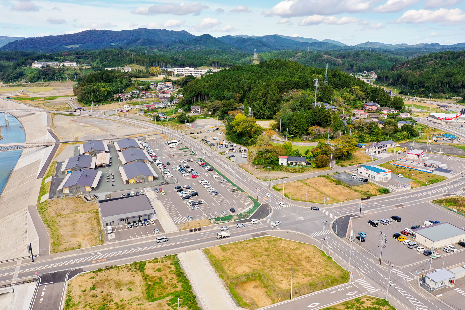 建物や駐車場、道路、緑地が広がり、複数の車が駐車や走行しており、背景には丘や山々が囲む様子が写された写真