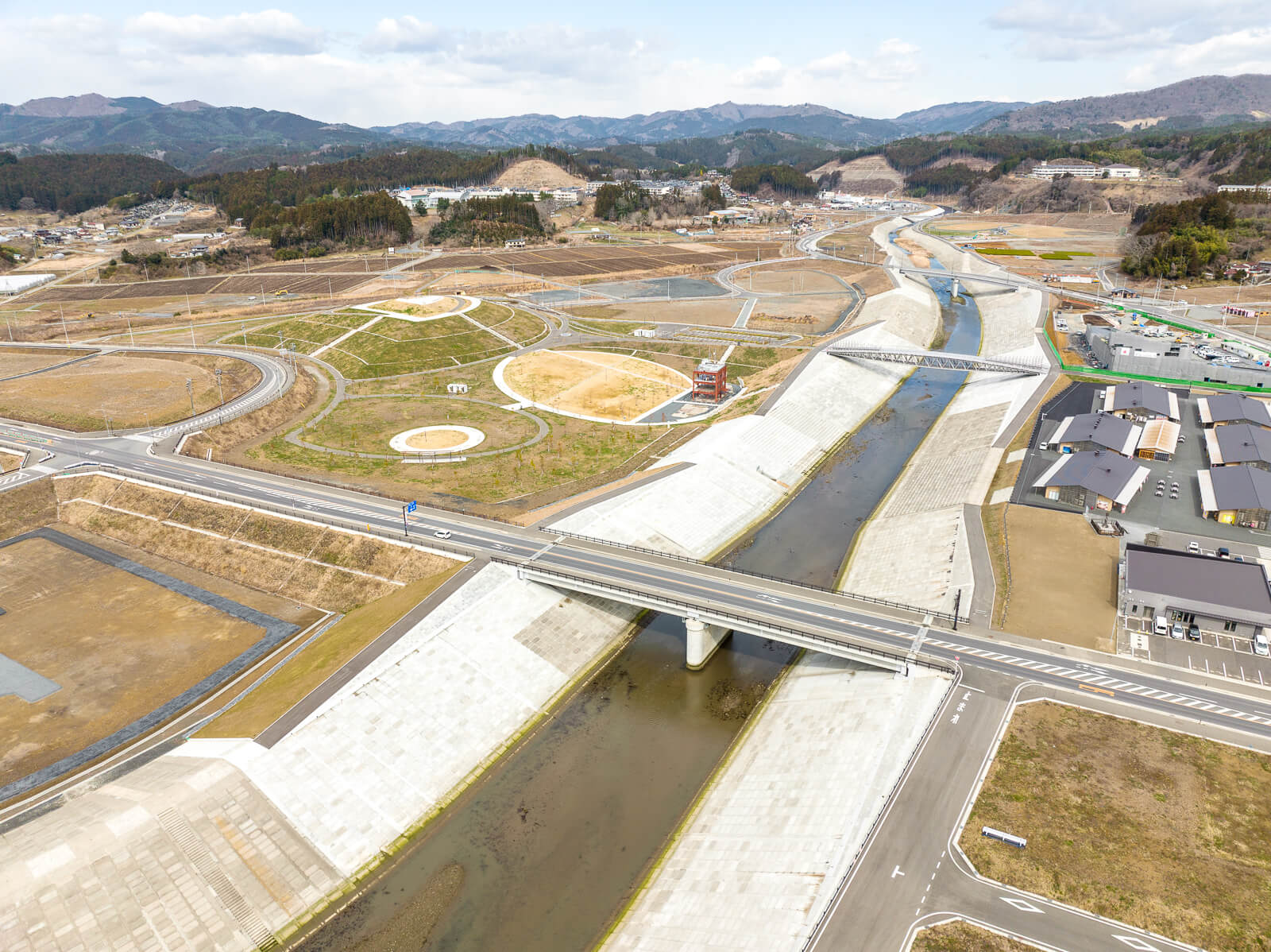 整備された川の両岸に道路と土地が整然と配置され、中央にある緑地には複数の円形の植栽スペースが見え、赤い建物も一つ立っており、背景には山が広がる復興した住宅予定地と公園の昼間の風景を高台から定点カメラで撮影した写真