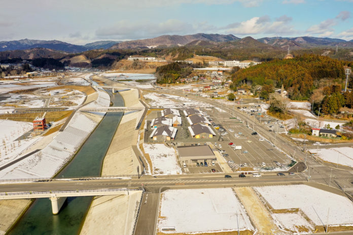中央に川が流れる風景を上空から撮影した画像で、川はコンクリートの護岸に沿って流れ、手前には橋が架かり、周囲には雪に覆われた土地や駐車場、複数の建物が点在し、背景には雪が残る丘や山が広がる冬の川沿い地域の写真