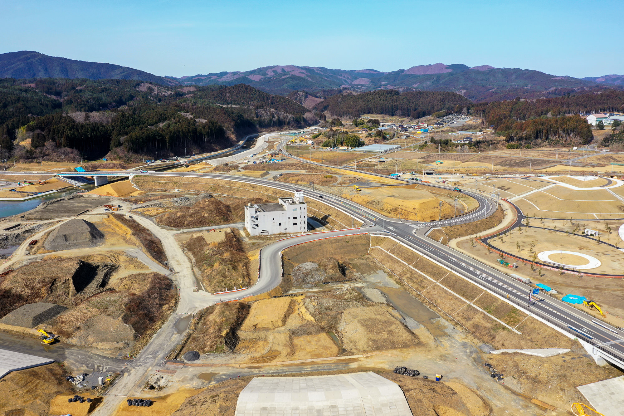 複数の道路と建物、重機が点在する広大な建設現場を空から見下ろし、周囲は起伏のある地形で一部が整地され、背景には山々が広がり、空は澄んだ青空が広がっている建設中の地域の写真
