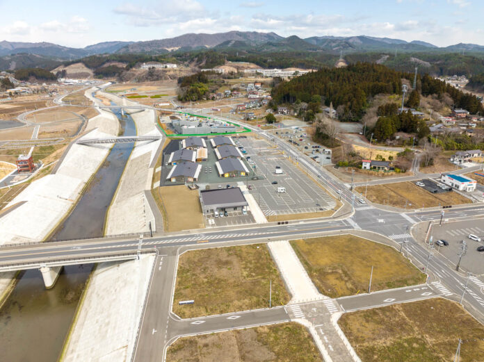 川に沿って整備された堤防と橋が見え、川の左側には広い駐車場と複数の平屋建ての建物が並ぶ商業施設があり、その背後には森林と住宅が広がっている、復興後の街の中心部の昼間の風景を高台から定点カメラで撮影した写真