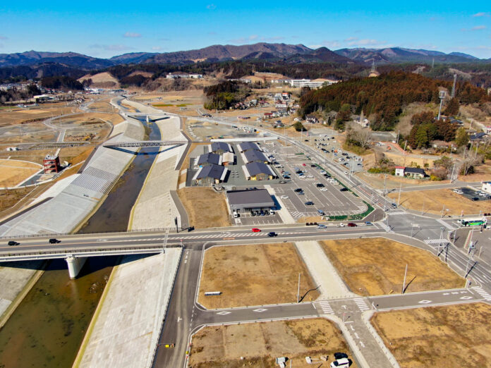 川が流れる開発済みの地域を上空から見下ろした風景が広がっており、川には橋が架かっていて、周囲には複数の建物や駐車場、道路や交差点が点在し、未開発の土地も一部見られ、背景には青空の下に広がる丘や山々が見える写真