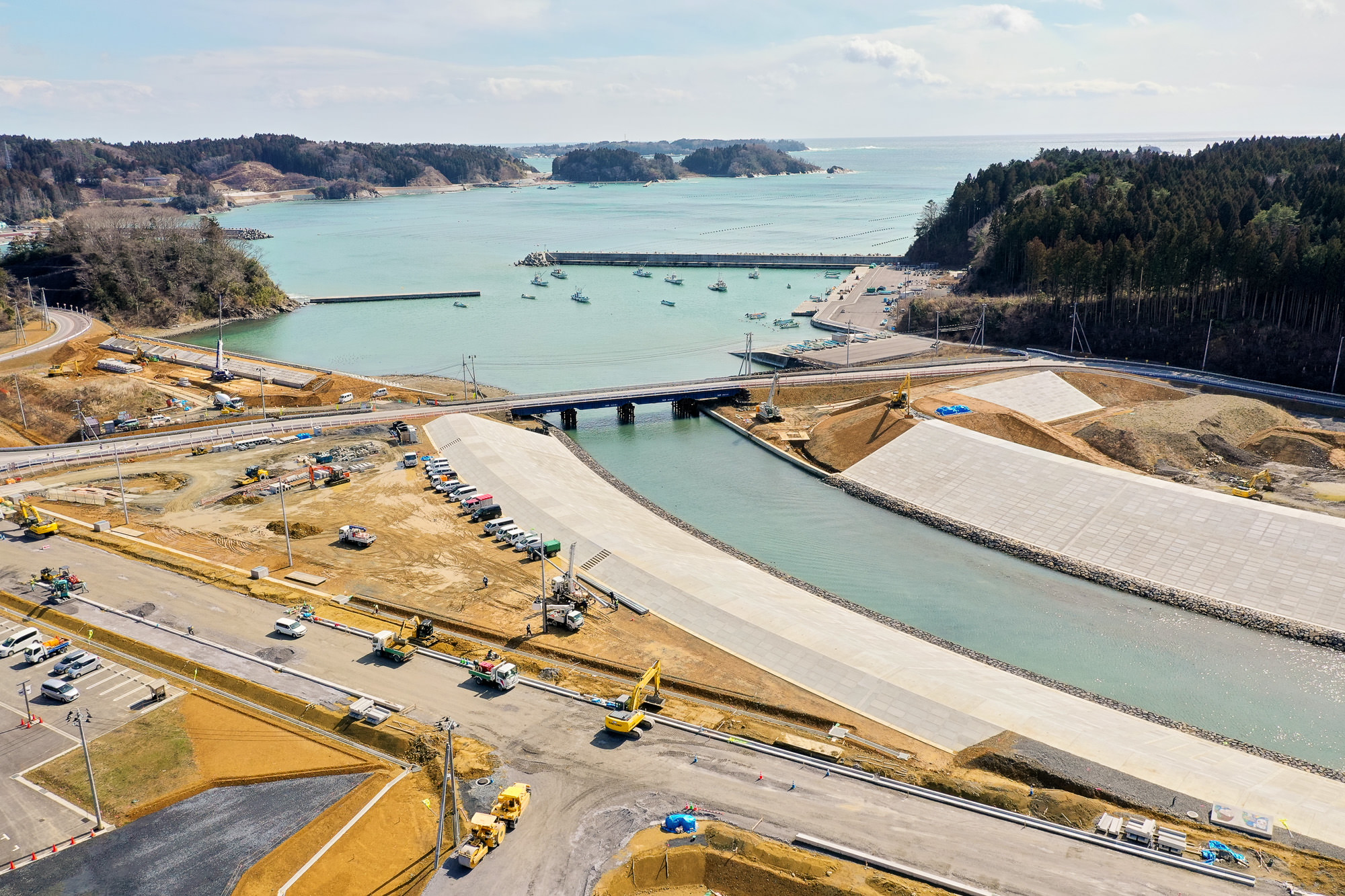 川が海へと流れ込む海岸沿いの工事現場を空から見下ろし、コンクリートの護岸や橋、複数の道路が整備され、クレーンやトラックなどの建設機材が点在し、背景には小型船が停泊する海と森林に覆われた丘が広がる海辺の工事現場の写真