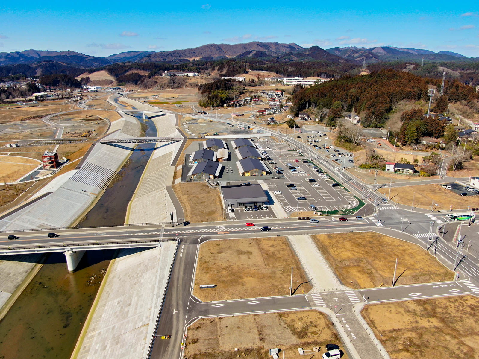 川が流れる開発済みの地域を上空から見下ろした風景が広がっており、川には複数の橋が架かり、周囲には道路や駐車場、建物が点在し、背景には丘や山々が広がっている写真