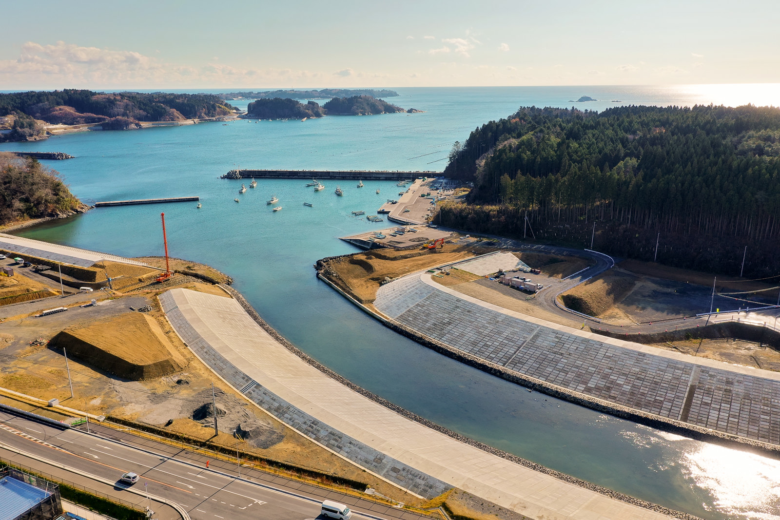 防波堤に囲まれた港を中心とした海岸地域を上空から撮影した風景で、港内には複数の船が停泊しており、周囲には道路やインフラが整備され、コンクリートで新しく造成されたと思われる区域も見られ、背景には森林や遠くに小さな島々が広がり、穏やかな海と晴れた空が広がる港湾地域の写真