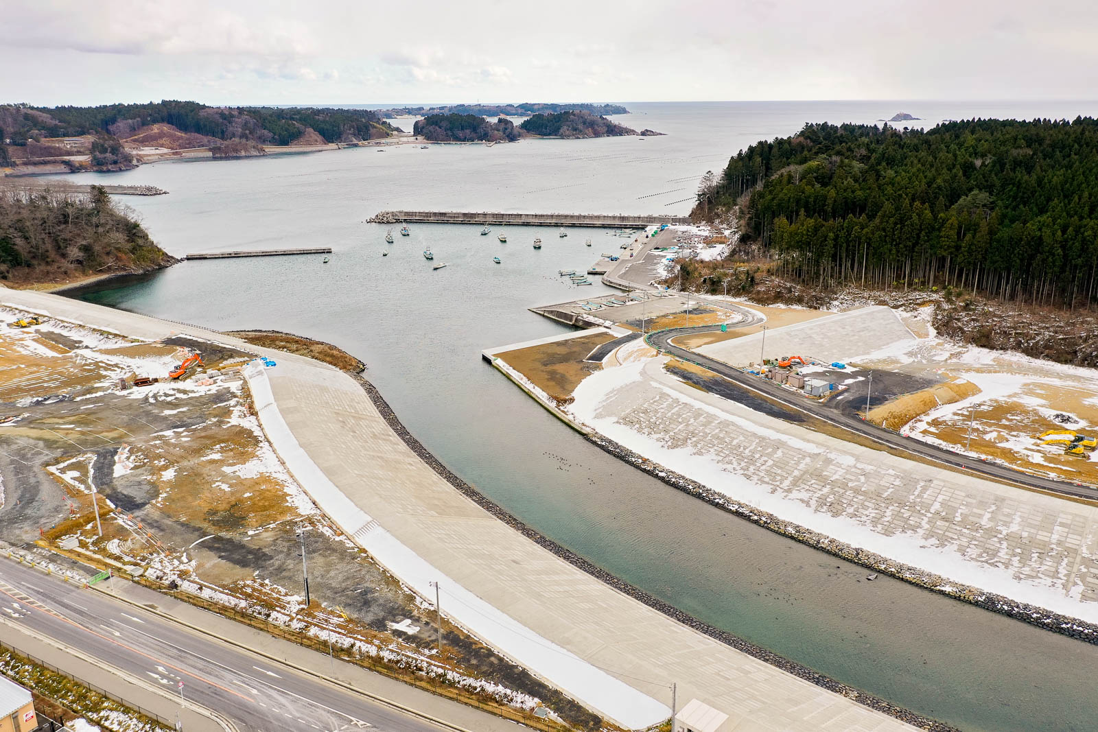 手前から奥に向かって緩やかにカーブする川の両岸がコンクリートで整備され、船が停泊している中央に防波堤があり、奥の林道には雪が積もっており杉林に囲まれた自然と人工構造物が混在する昼間の風景を高台から定点カメラで撮影した写真