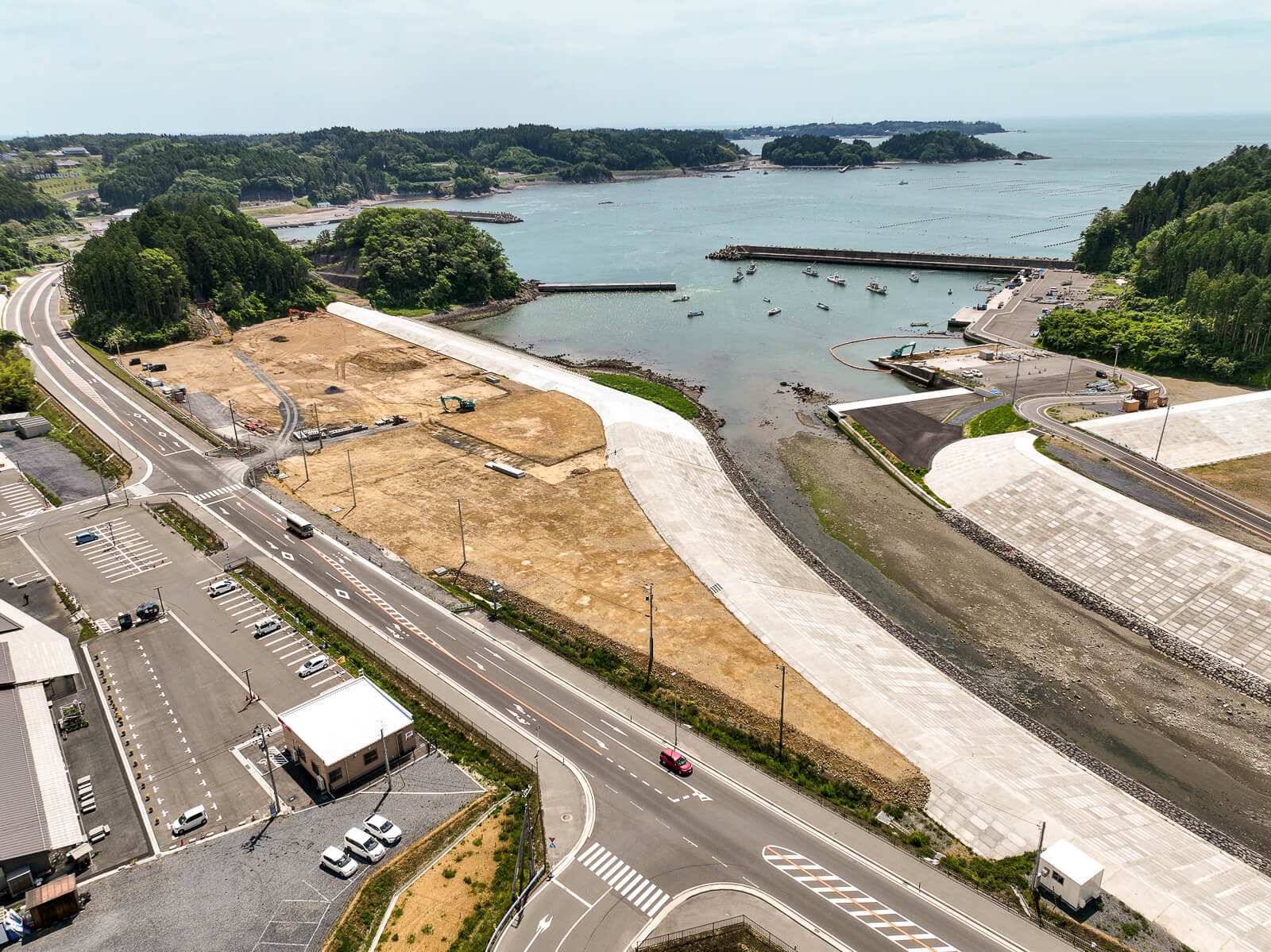 広い防波堤が海に向かって延びており、その周囲に未舗装の土地と工事用重機が点在し、奥に海と小さな船が停泊する港が見える昼間の風景を高台から定点カメラで撮影した写真