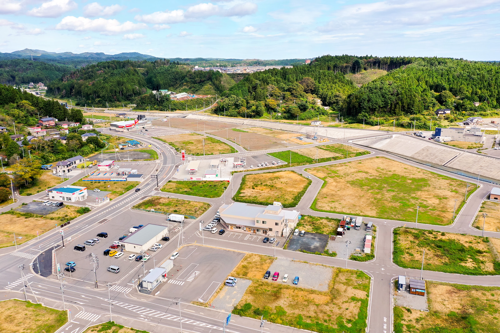 空から見下ろした風景で、建物や道路、駐車場が点在し、間には緑の草地が広がり、背景には森林に覆われた丘と遠くの山々が見える風景写真
